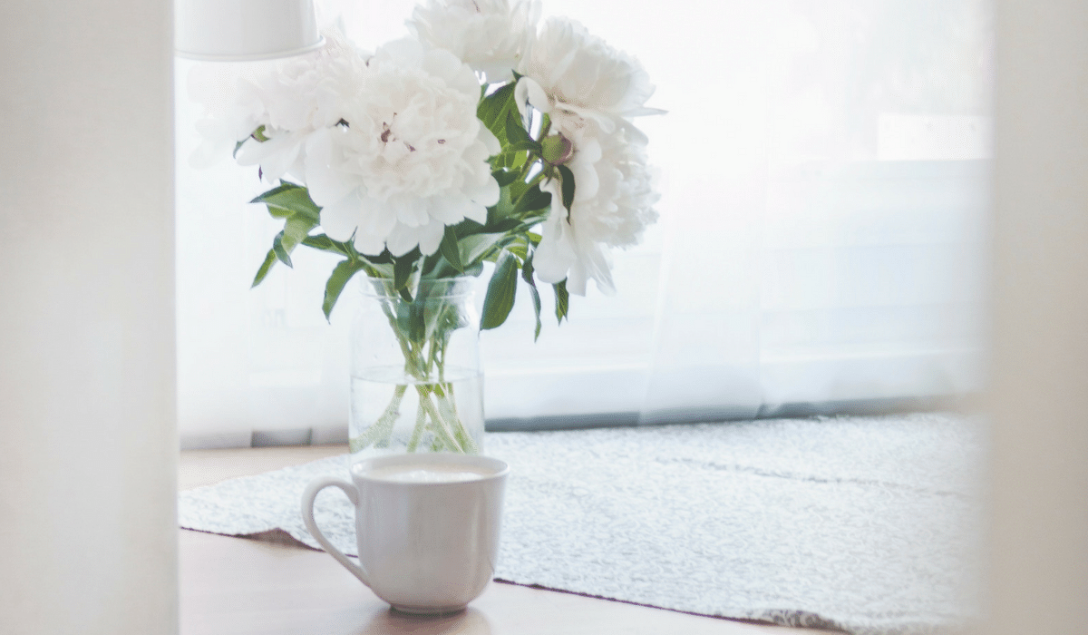 A cup of milk and a vase of white flowers on a table.