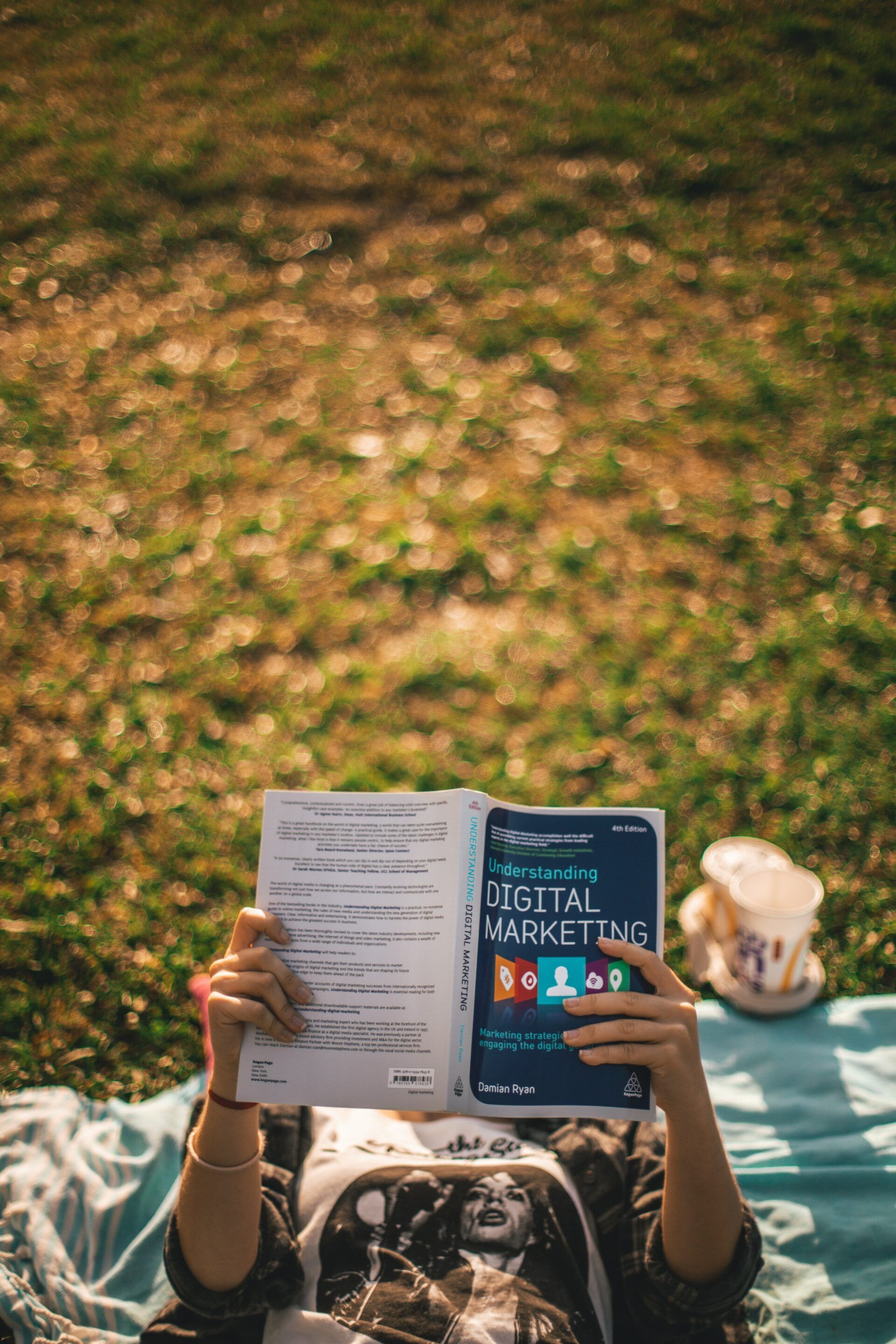 A person is laying on a blanket in the grass reading a book.