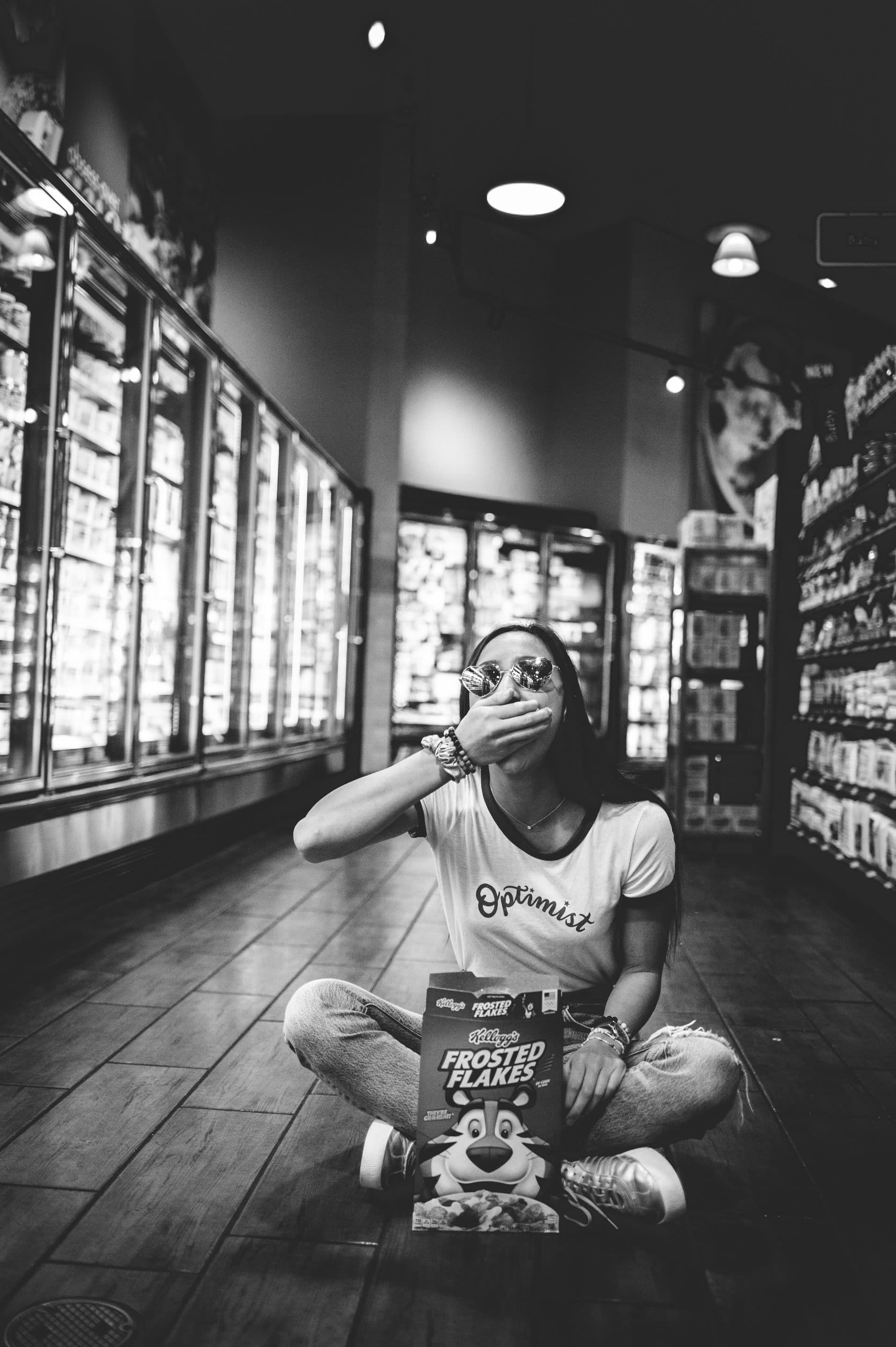 A woman is sitting on the floor in a grocery store eating a box of cereal.