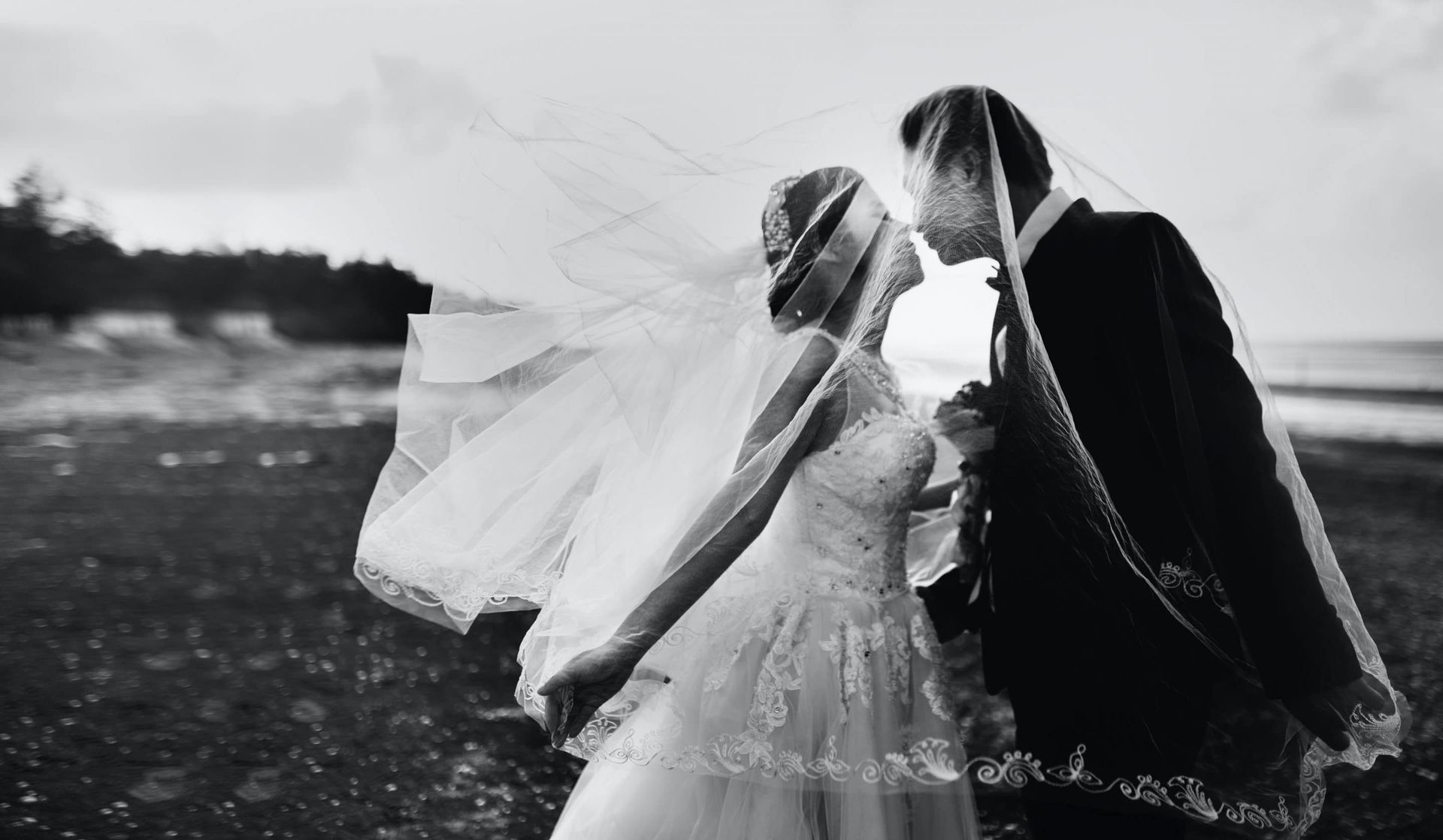 A bride and groom kissing under a veil on a beach.