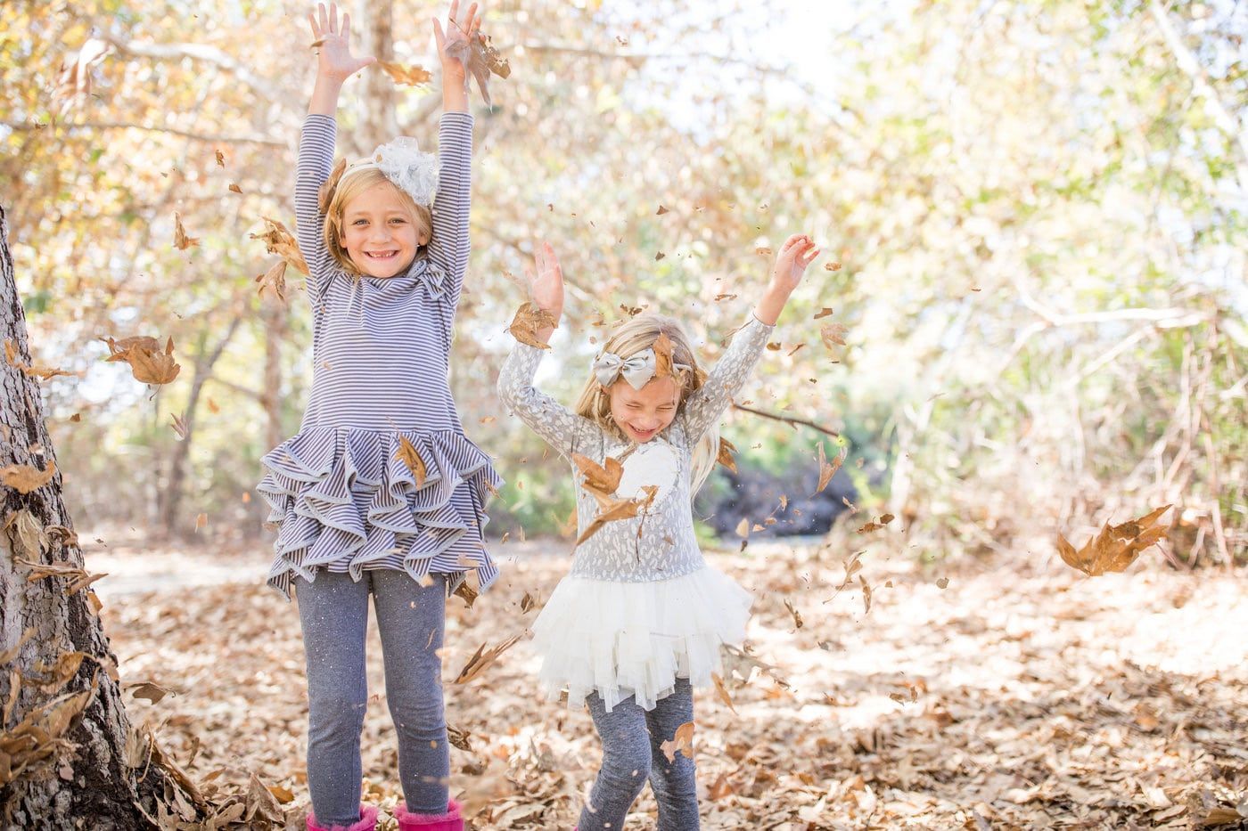 Two little girls are throwing leaves in the air in a park.