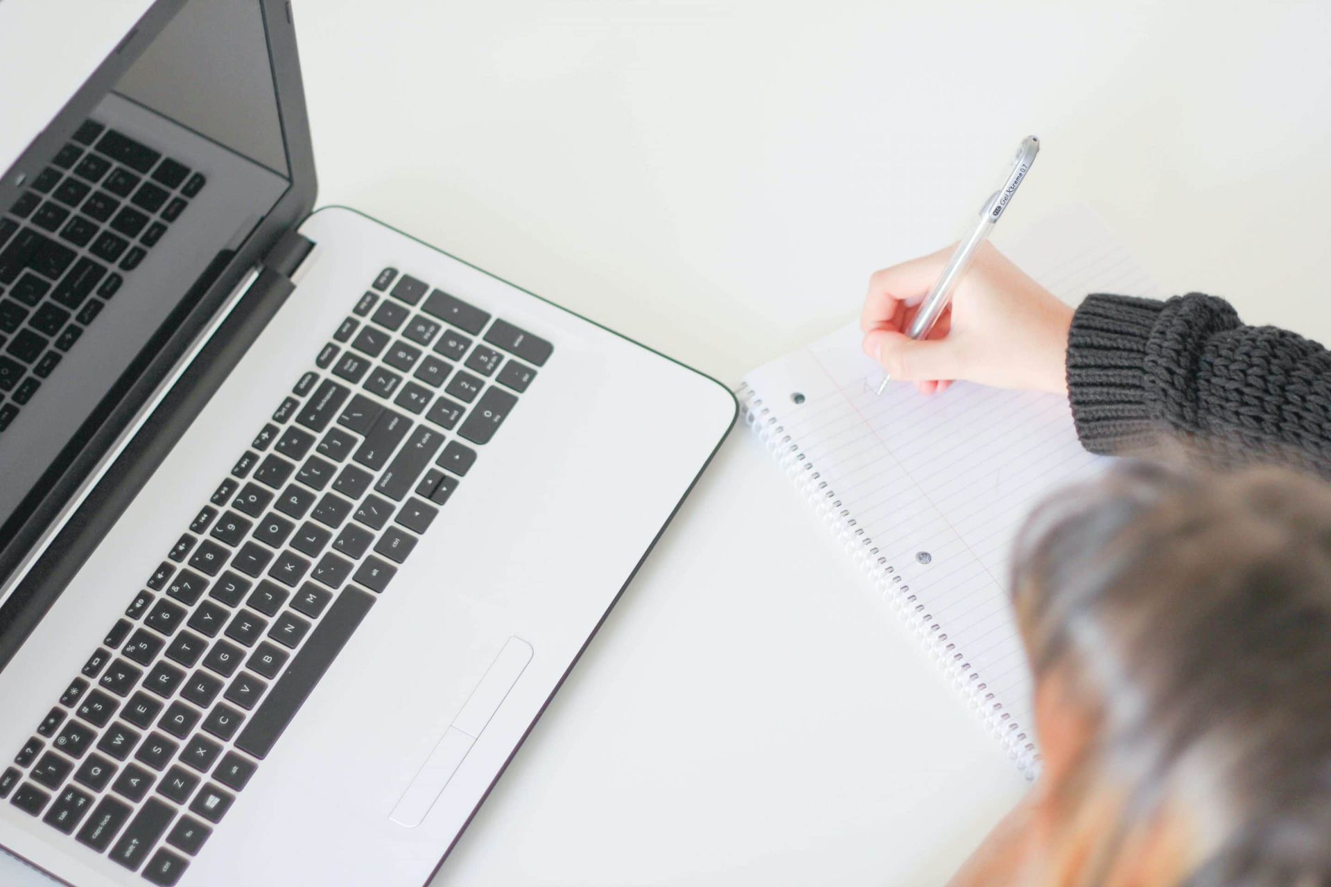 A person is writing in a notebook in front of a laptop computer.