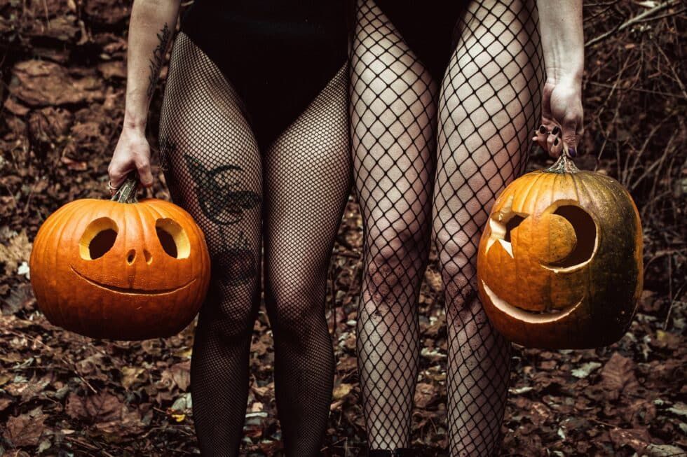 Two women in fishnet stockings are holding carved pumpkins.