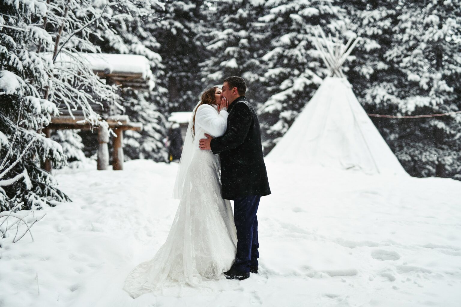 A bride and groom are kissing in the snow in front of a teepee.