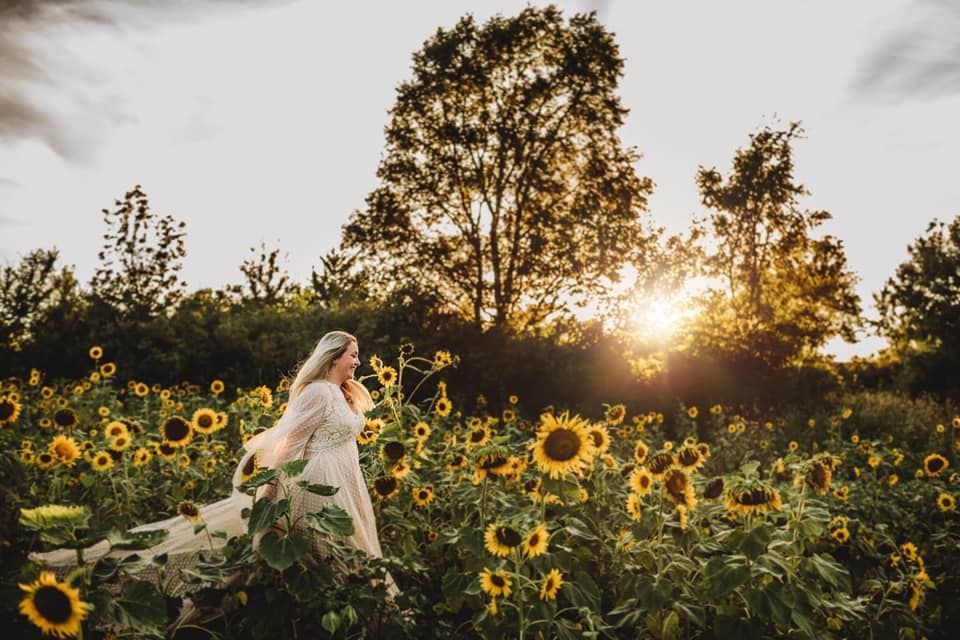 A bride is standing in a field of sunflowers at sunset.