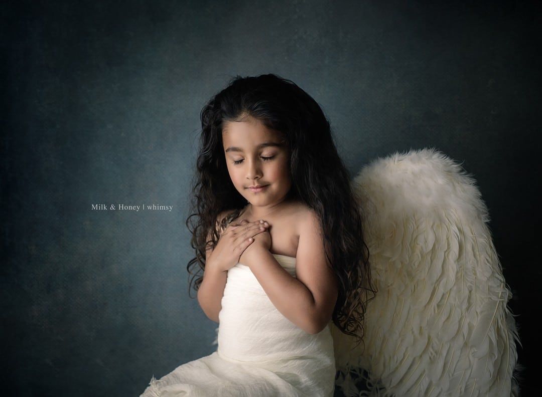 A little girl wearing angel wings is sitting on a chair.