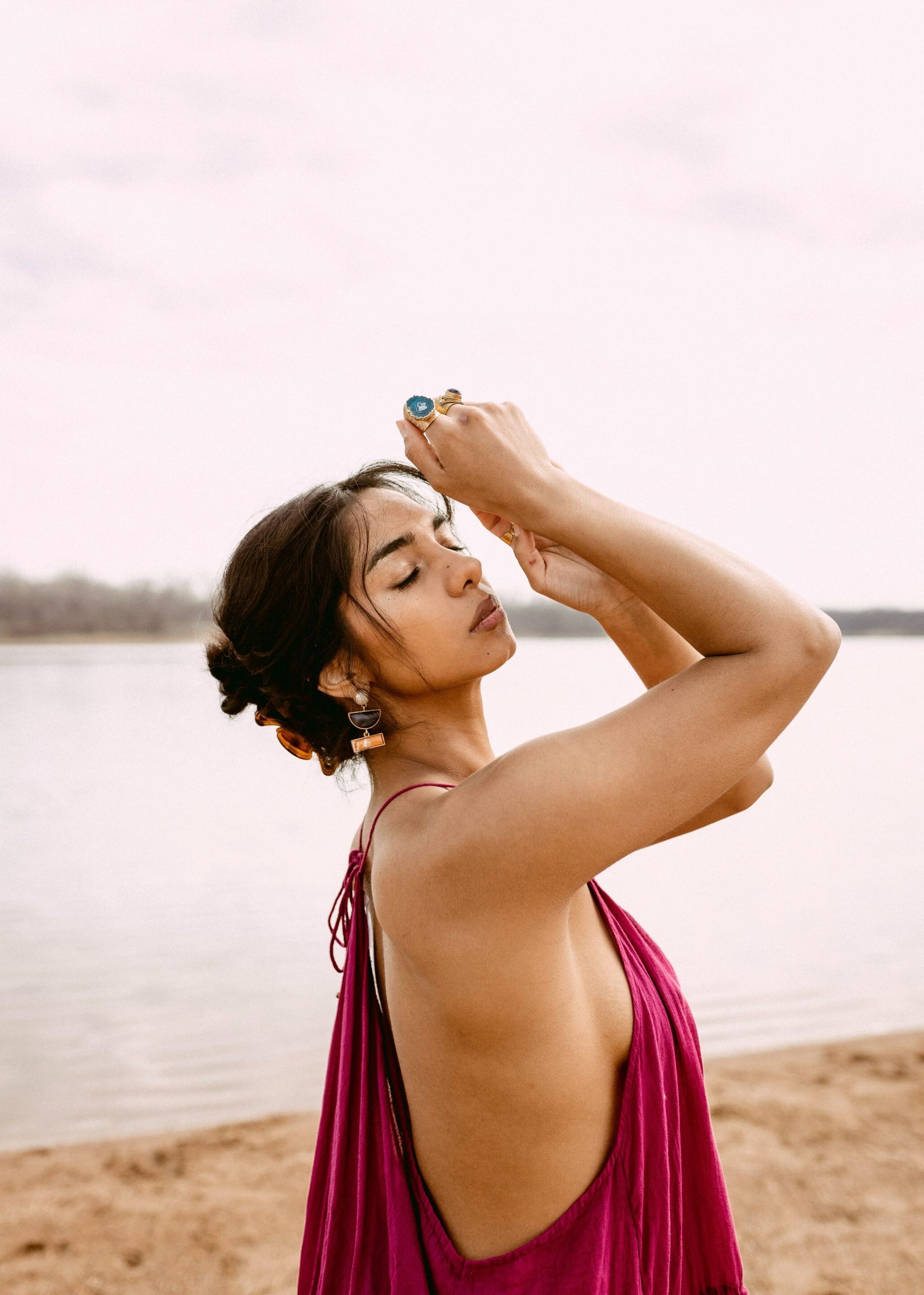 A woman in a pink dress is standing on a beach with her hands on her head.