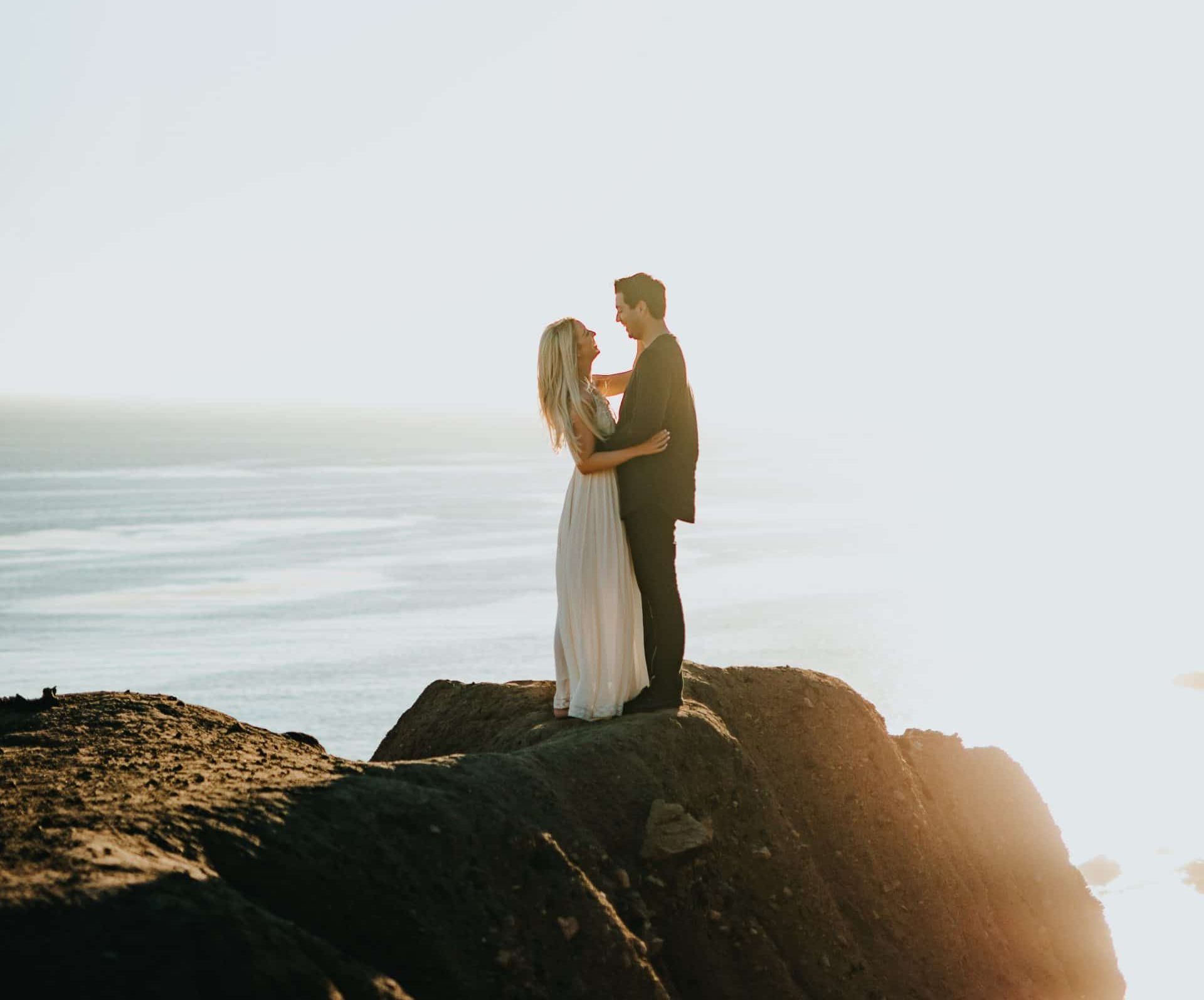 A bride and groom are standing on top of a cliff overlooking the ocean.