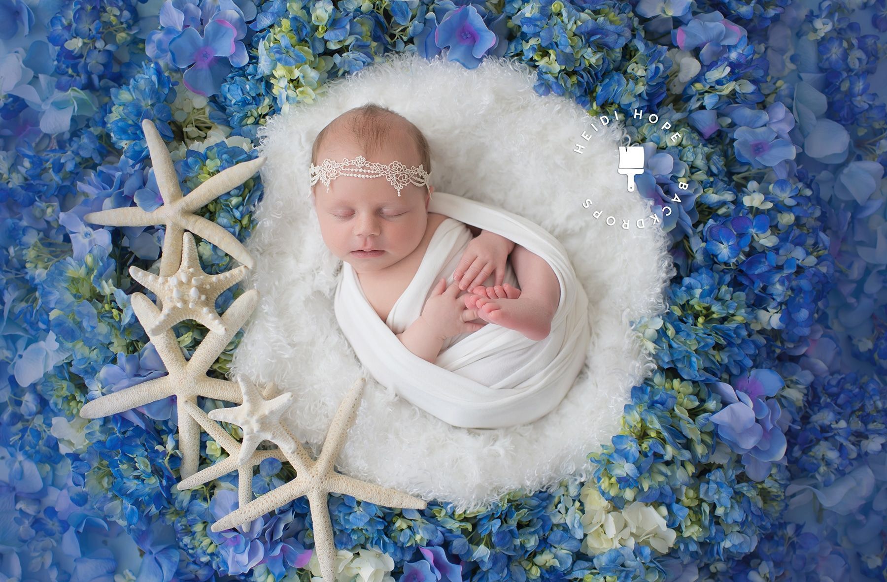 A newborn baby is wrapped in a white blanket surrounded by blue flowers and starfish.