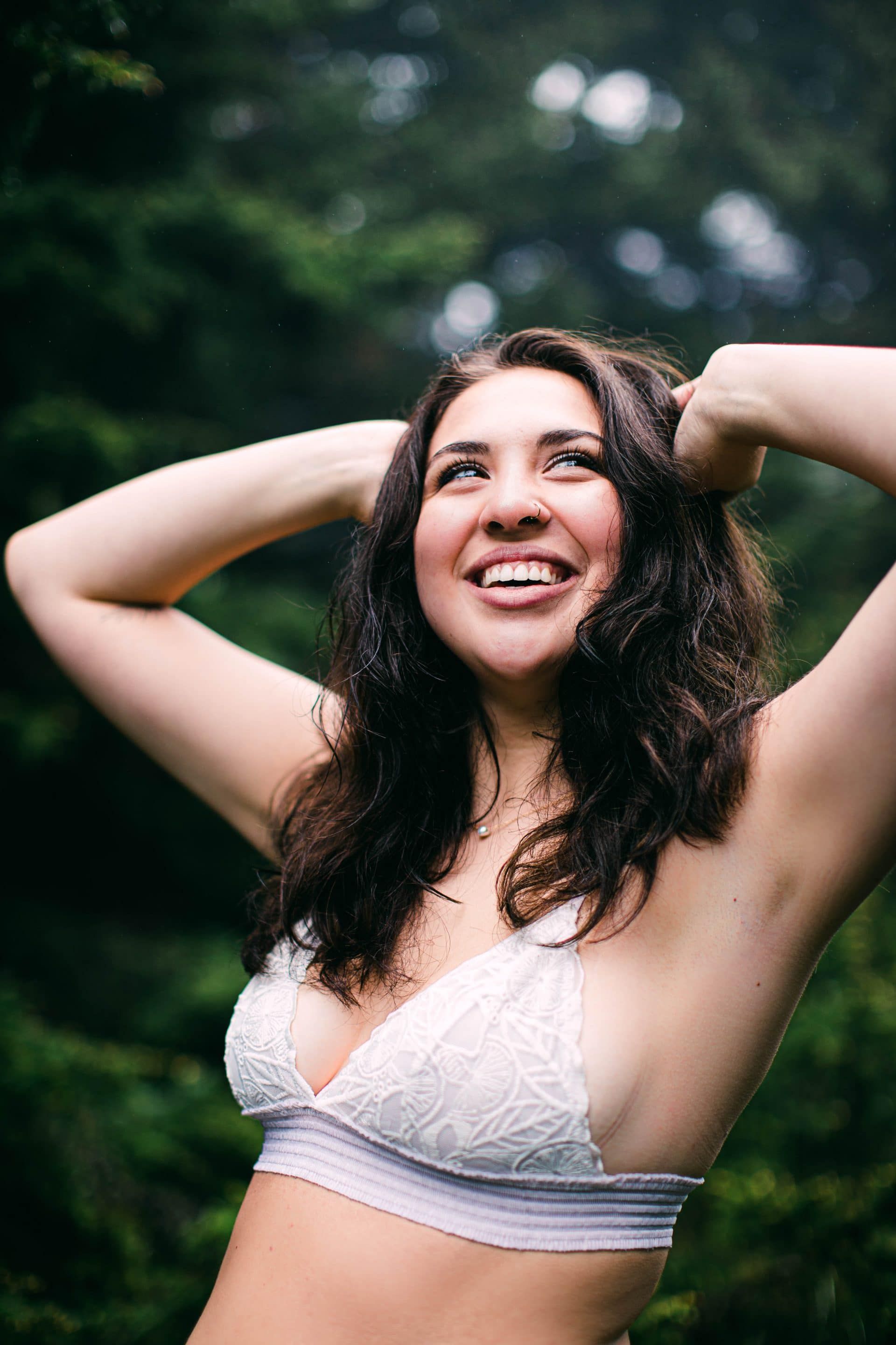 A woman in a white bra is standing in the woods with her hands in her hair.