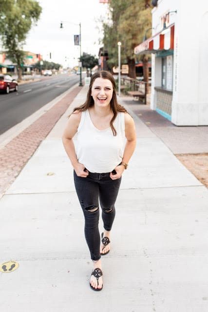 A woman in a white tank top and black jeans is walking down a sidewalk.