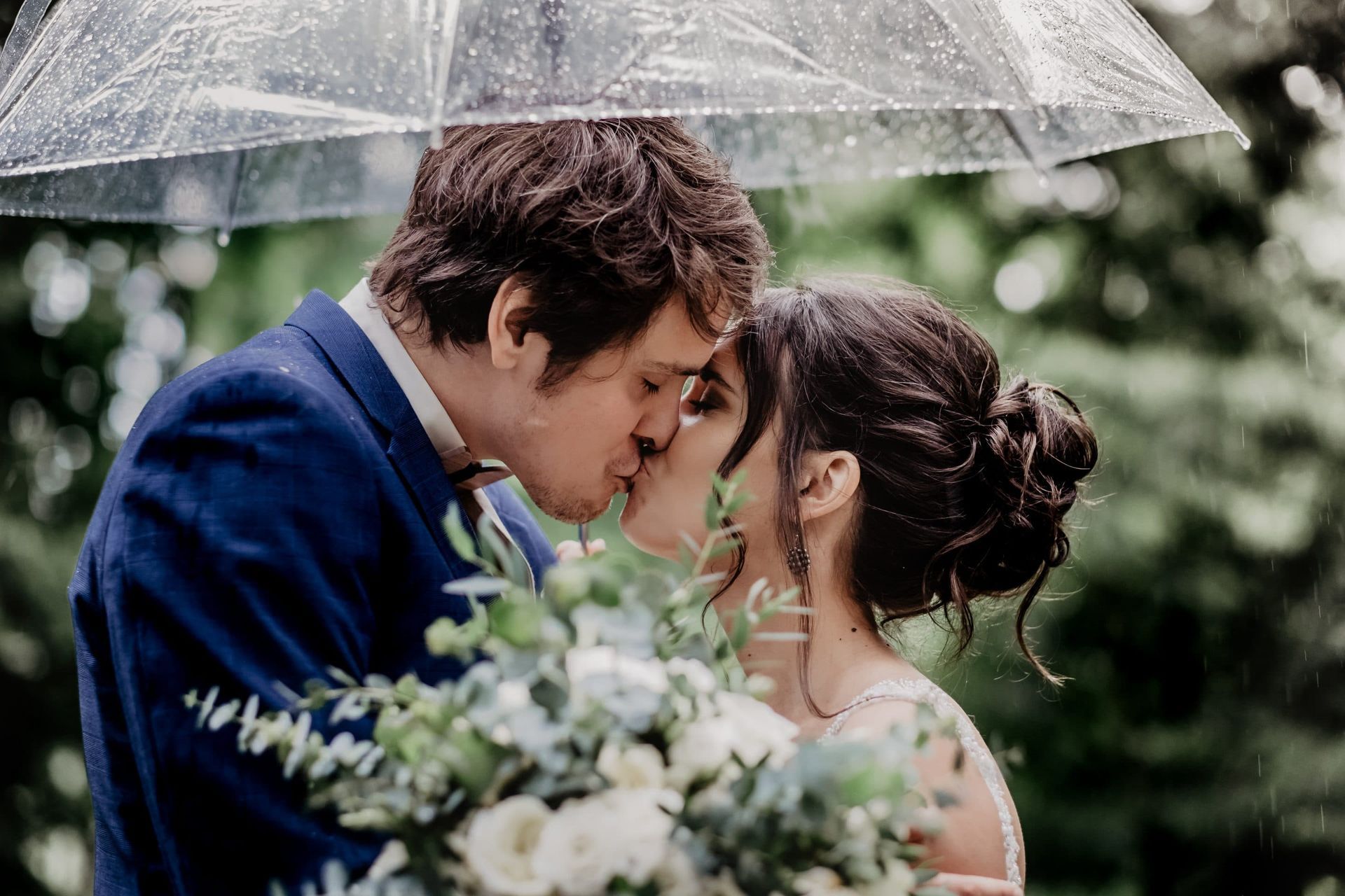 A bride and groom are kissing under an umbrella in the rain.