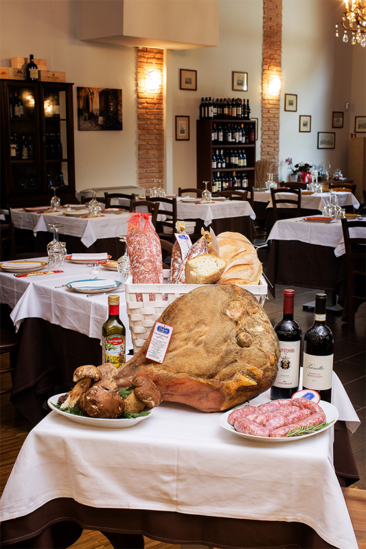 Tavolo con esposizione di prodotti alimentari italiani: prosciutto stagionato, salsicce, pane, bottiglie di vino e ambientazione da ristorante.