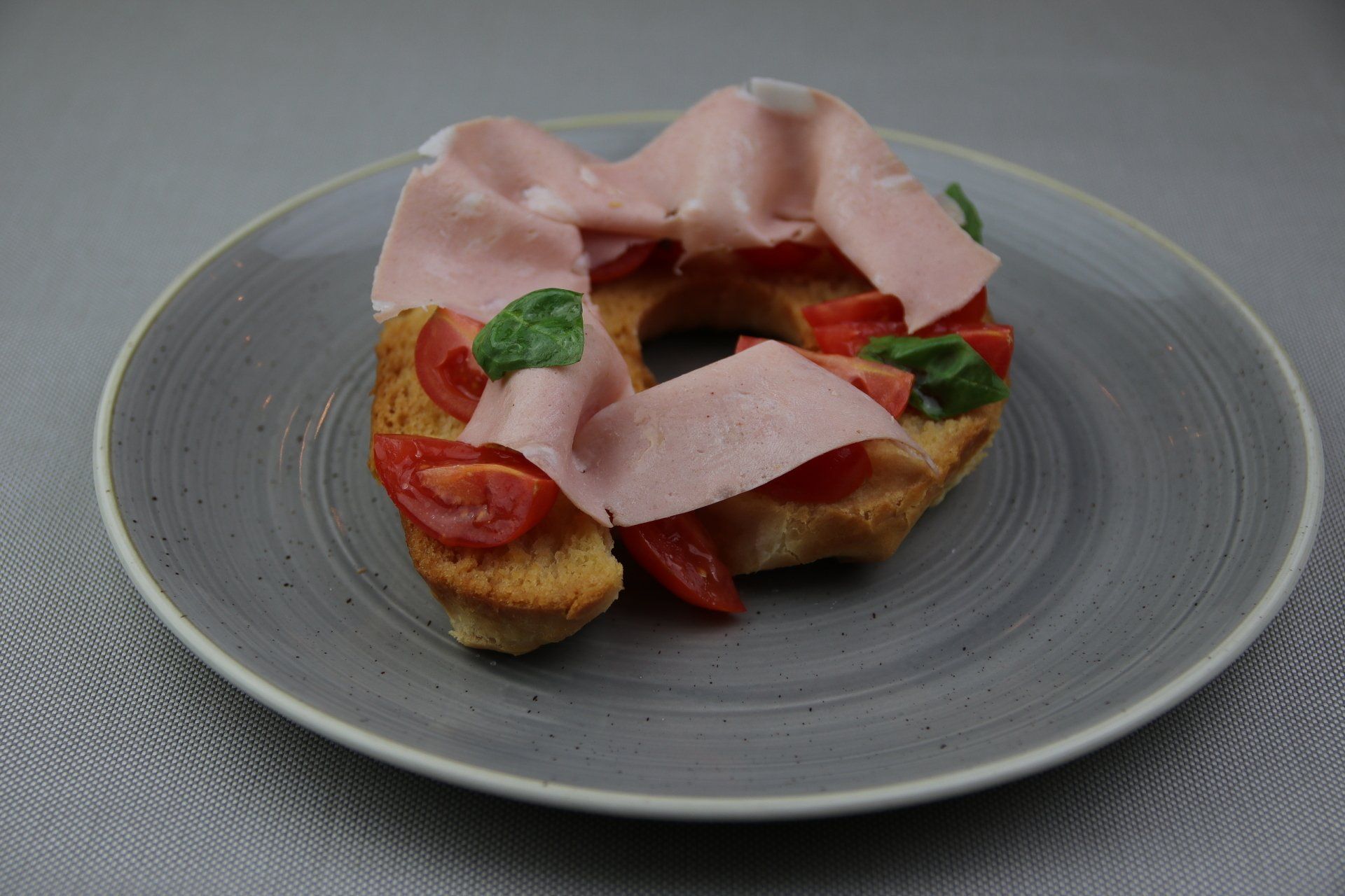 Pane tostato con mortadella, pomodori e basilico su un piatto grigio.