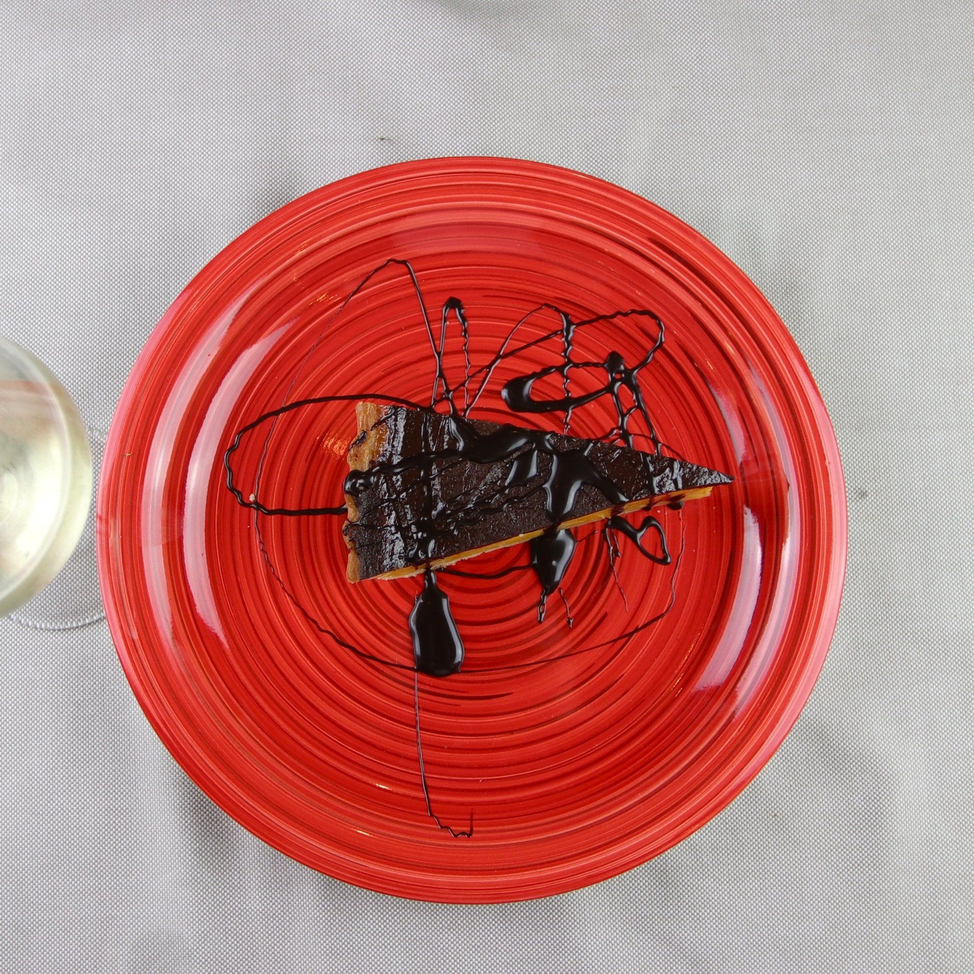 Fetta di torta al cioccolato su un piatto rosso, ricoperta di cioccolato, su una tovaglia grigia.