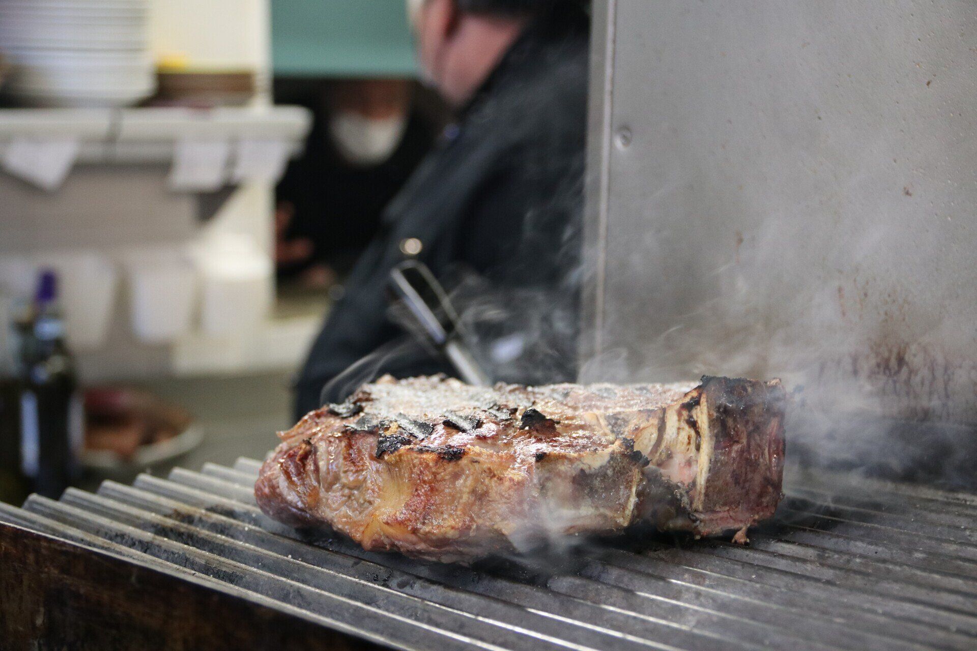 Una grande bistecca alla fiorentina viene cotta su una griglia metallica, con il fumo che sale. Sullo sfondo c'è uno chef in nero.