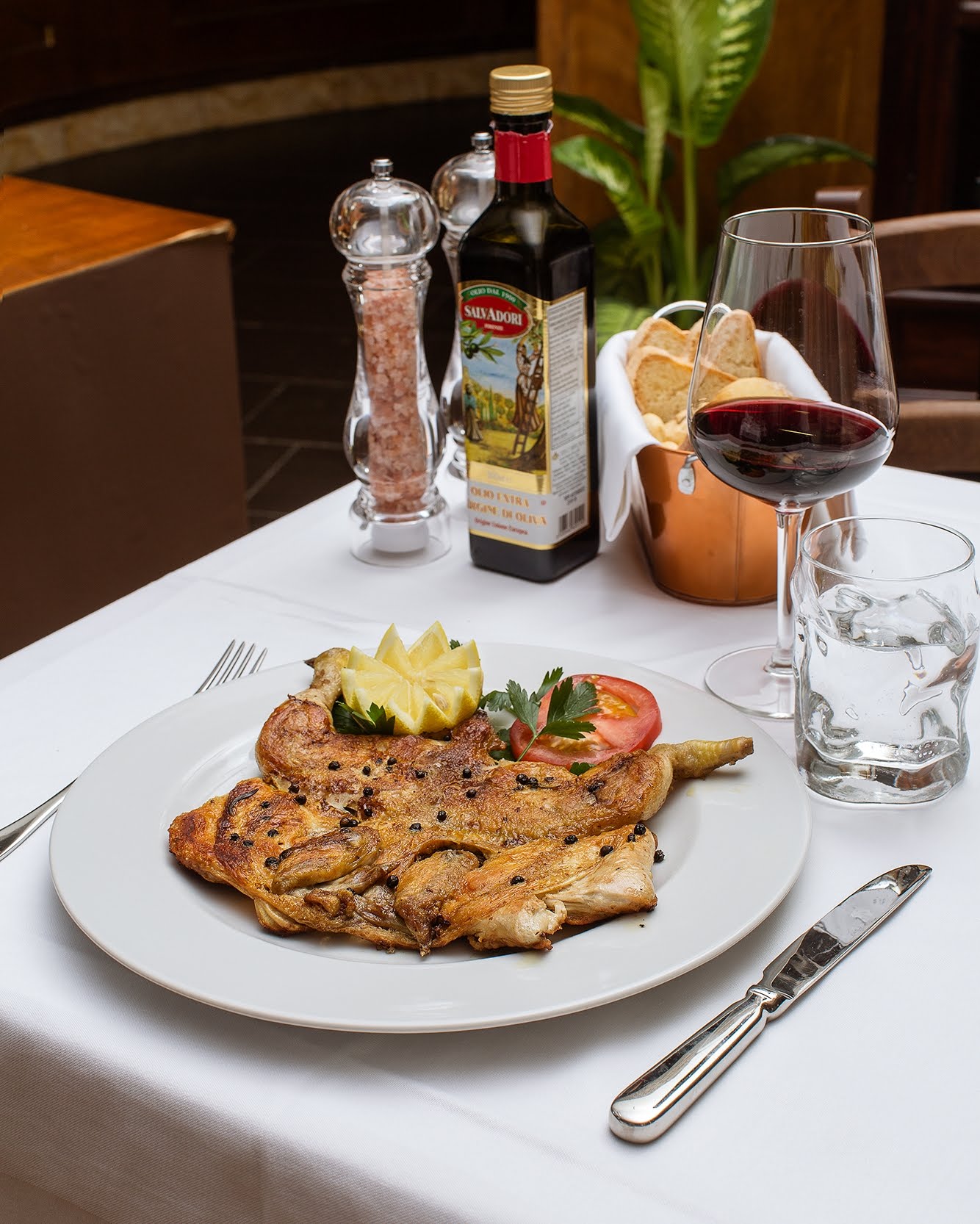 Pollo grigliato su un piatto bianco, apparecchiato a tavola con sale, olio, vino e pane.