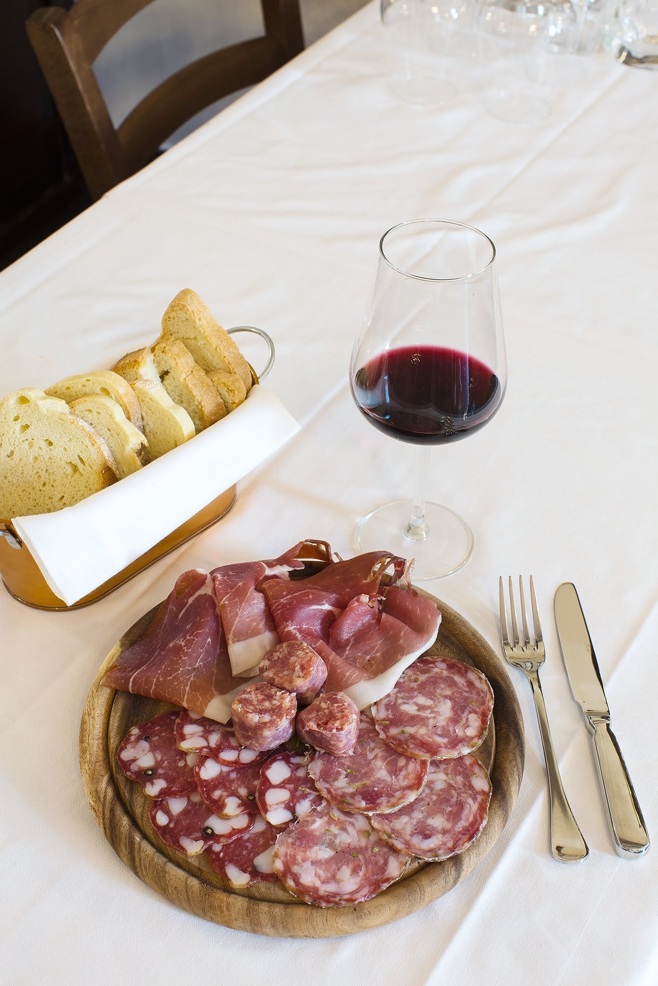 Piatto di salumi con pane, vino e posate su una tovaglia bianca.