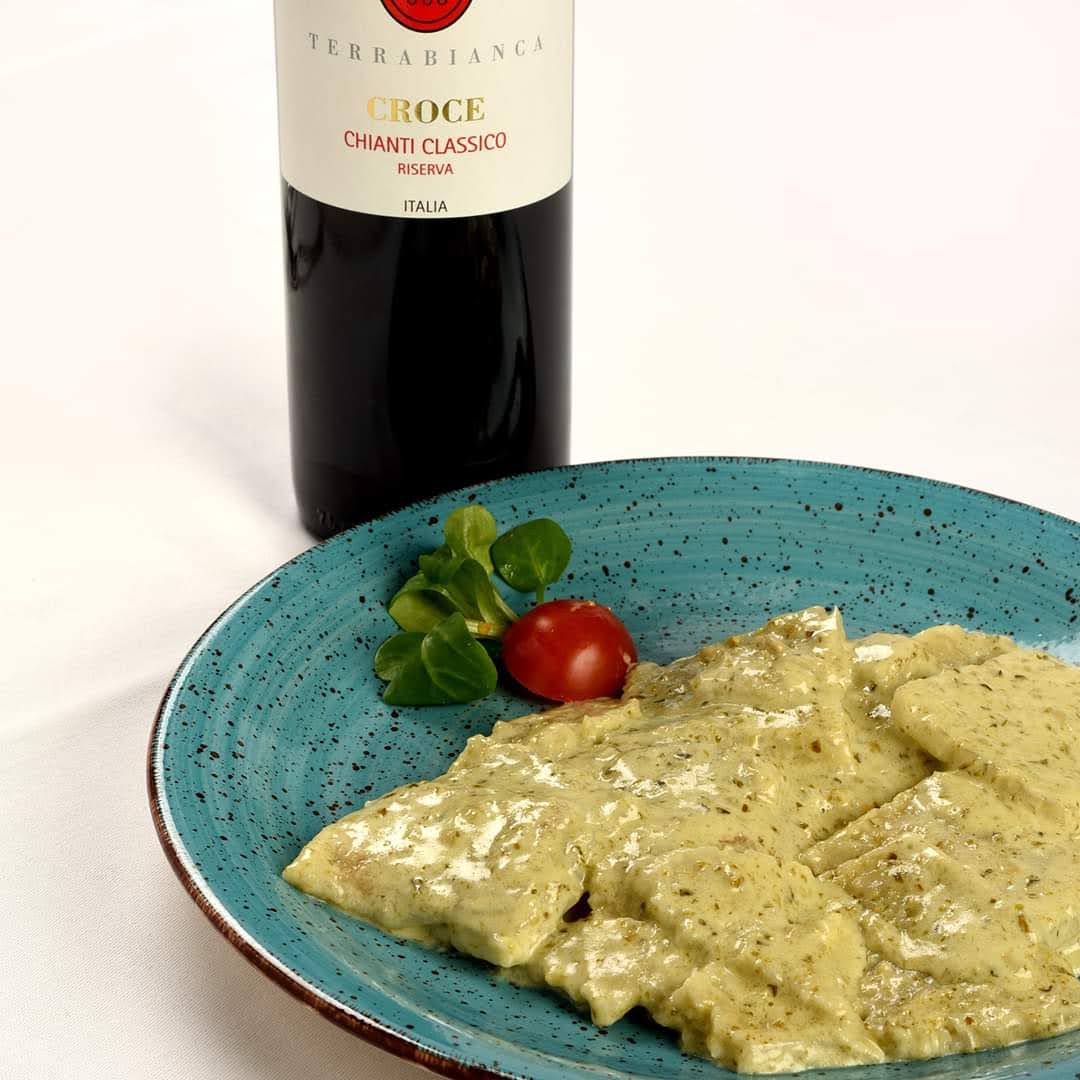 Ravioli con salsa cremosa, pomodorini e bottiglia di vino su un piatto blu, sfondo di tovaglia bianca.