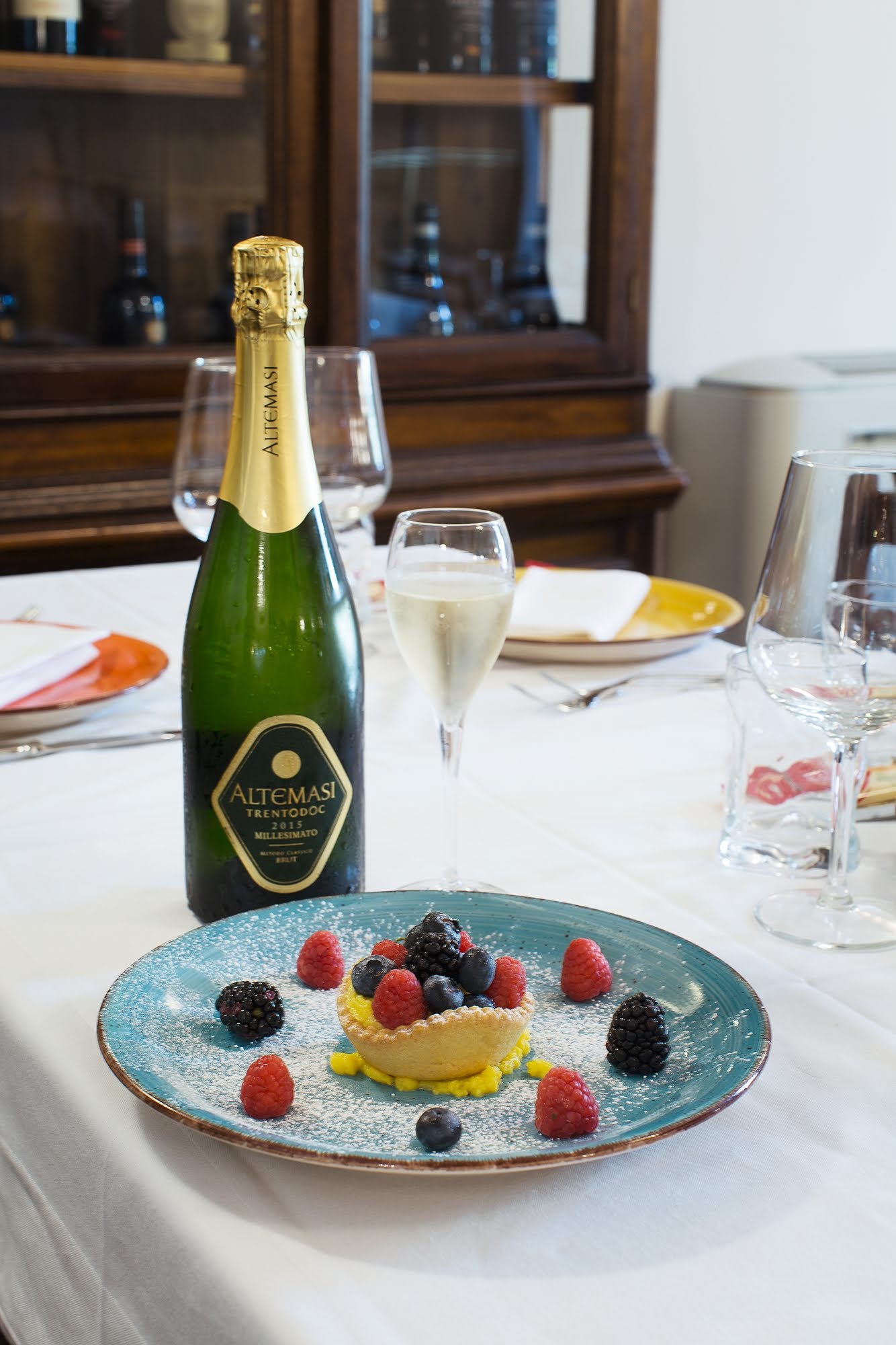 Una bottiglia di spumante e un dessert con frutti di bosco su una tavola apparecchiata per cenare.