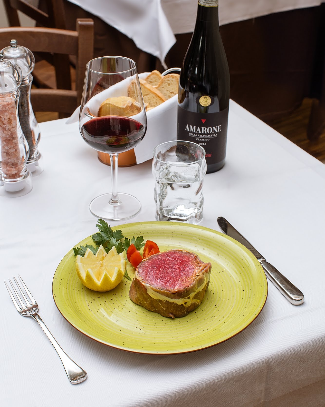 Tavolo da pranzo apparecchiato con cibo, vino, acqua e una bistecca su un piatto verde, tovaglia bianca, ristorante.