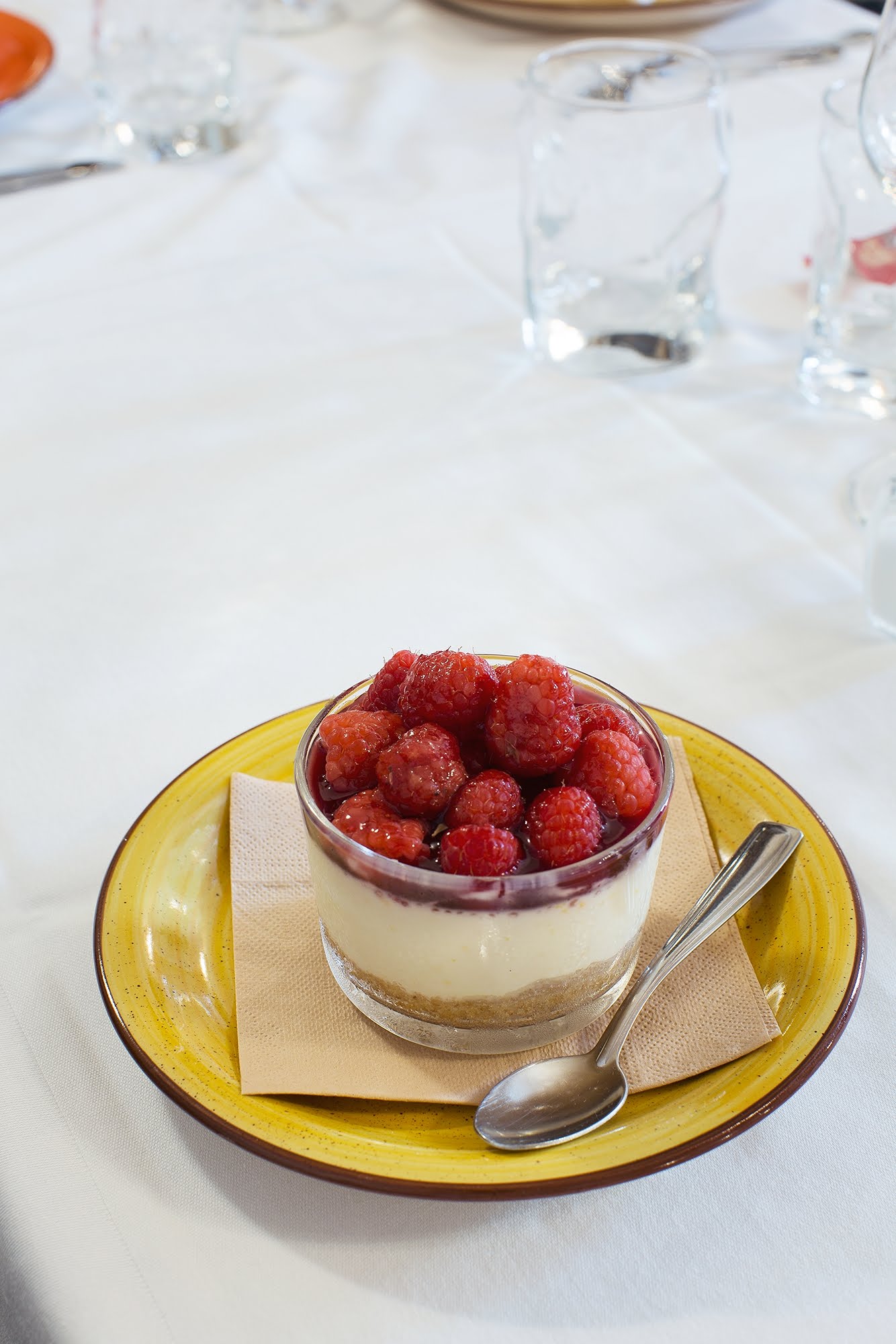 Cheesecake con lamponi servita su un piatto giallo, accanto a un cucchiaio, su una tovaglia bianca.
