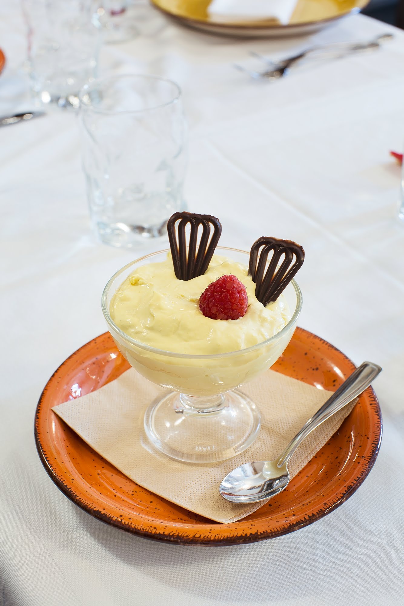 Dessert in una coppetta di vetro con guarnizioni di lampone e cioccolato su un piatto arancione, tovaglia bianca.