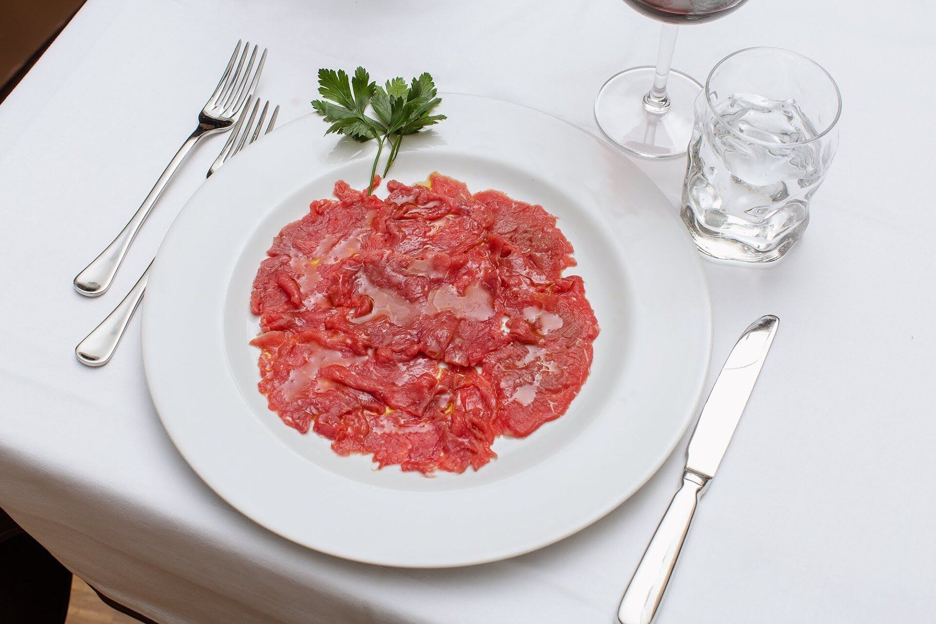 Carpaccio su un piatto bianco con posate e un bicchiere d'acqua, il tutto adagiato su una tovaglia bianca.