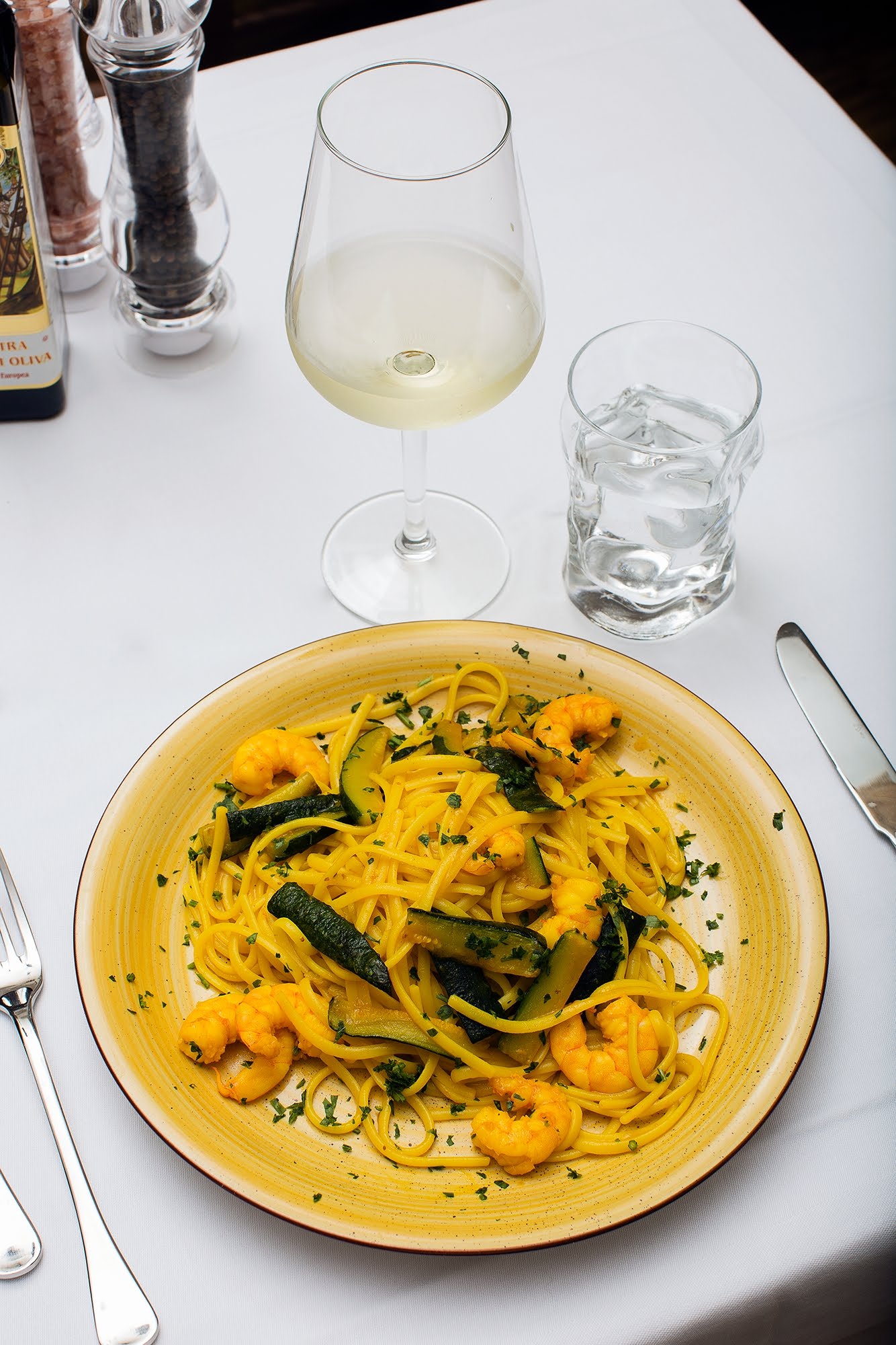Spaghetti con gamberi e zucchine su un piatto, con un bicchiere di vino bianco e acqua su una tovaglia bianca.