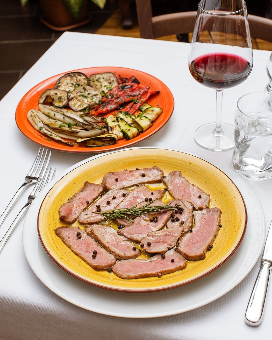 Verdure grigliate e carne affettata su piatti, bicchiere di vino, posate su una tovaglia bianca.