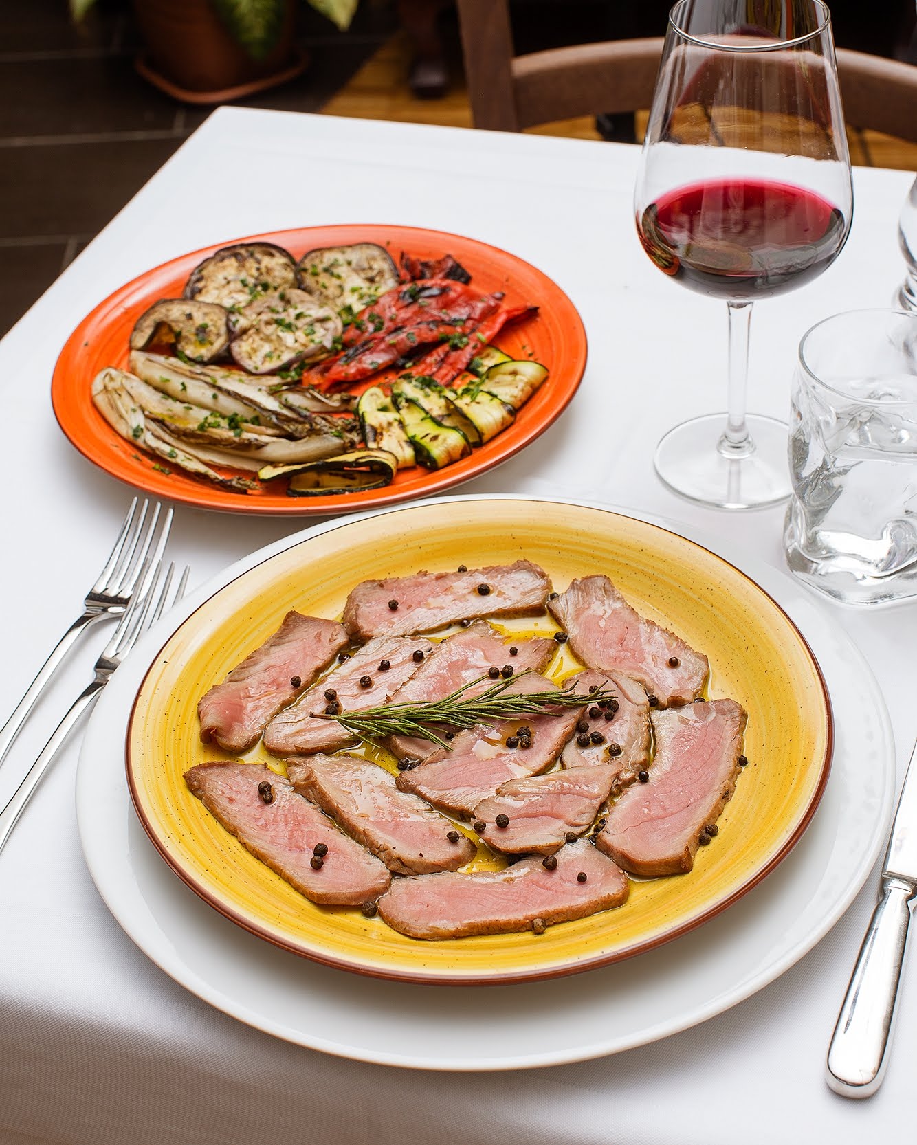 Piatto di carne affettata con grani di pepe e verdure grigliate su un tavolo apparecchiato con vino.