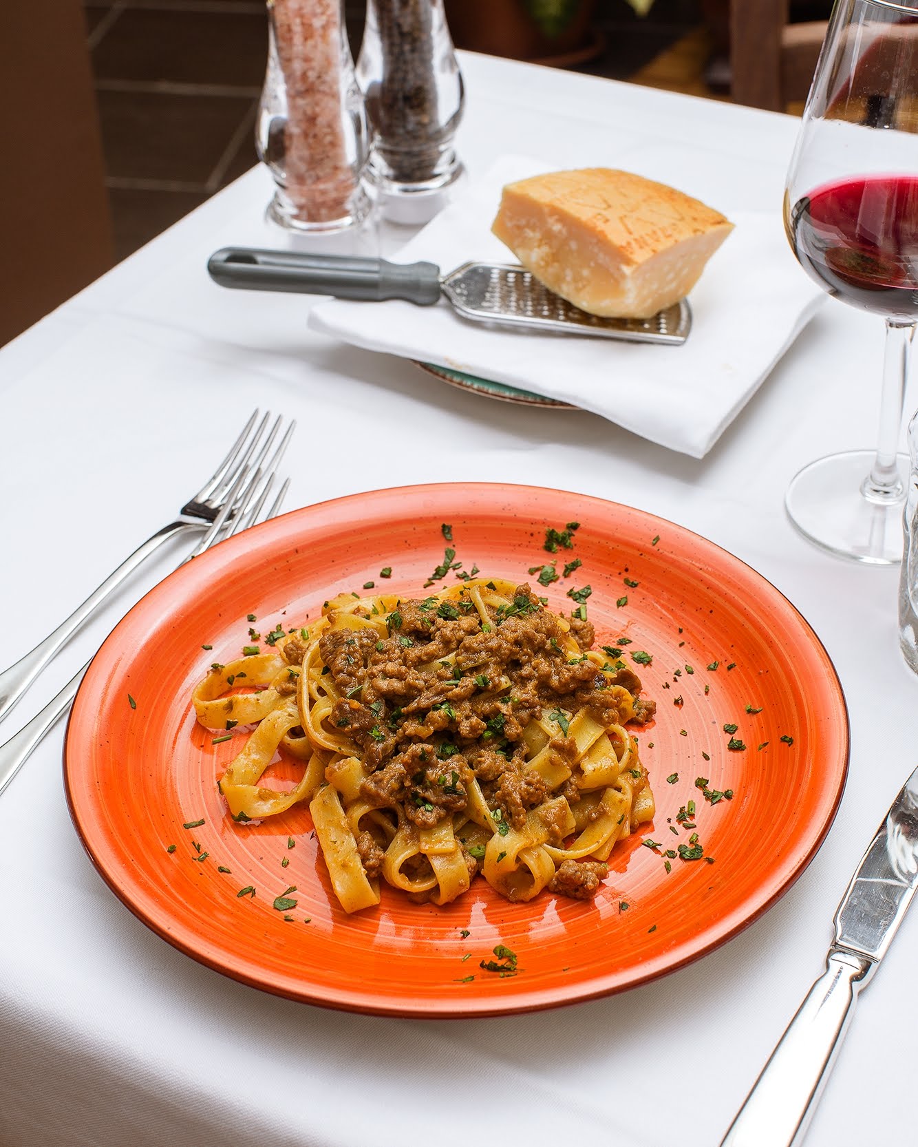 Pasta alla bolognese su un piatto arancione, con parmigiano, vino e posate su una tovaglia bianca.