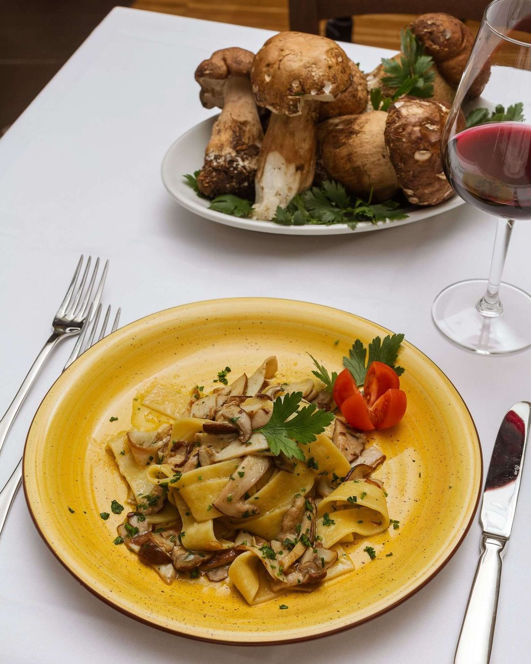 Pasta con funghi su un piatto giallo, funghi su un piatto, bicchiere di vino, posate su una tovaglia bianca.