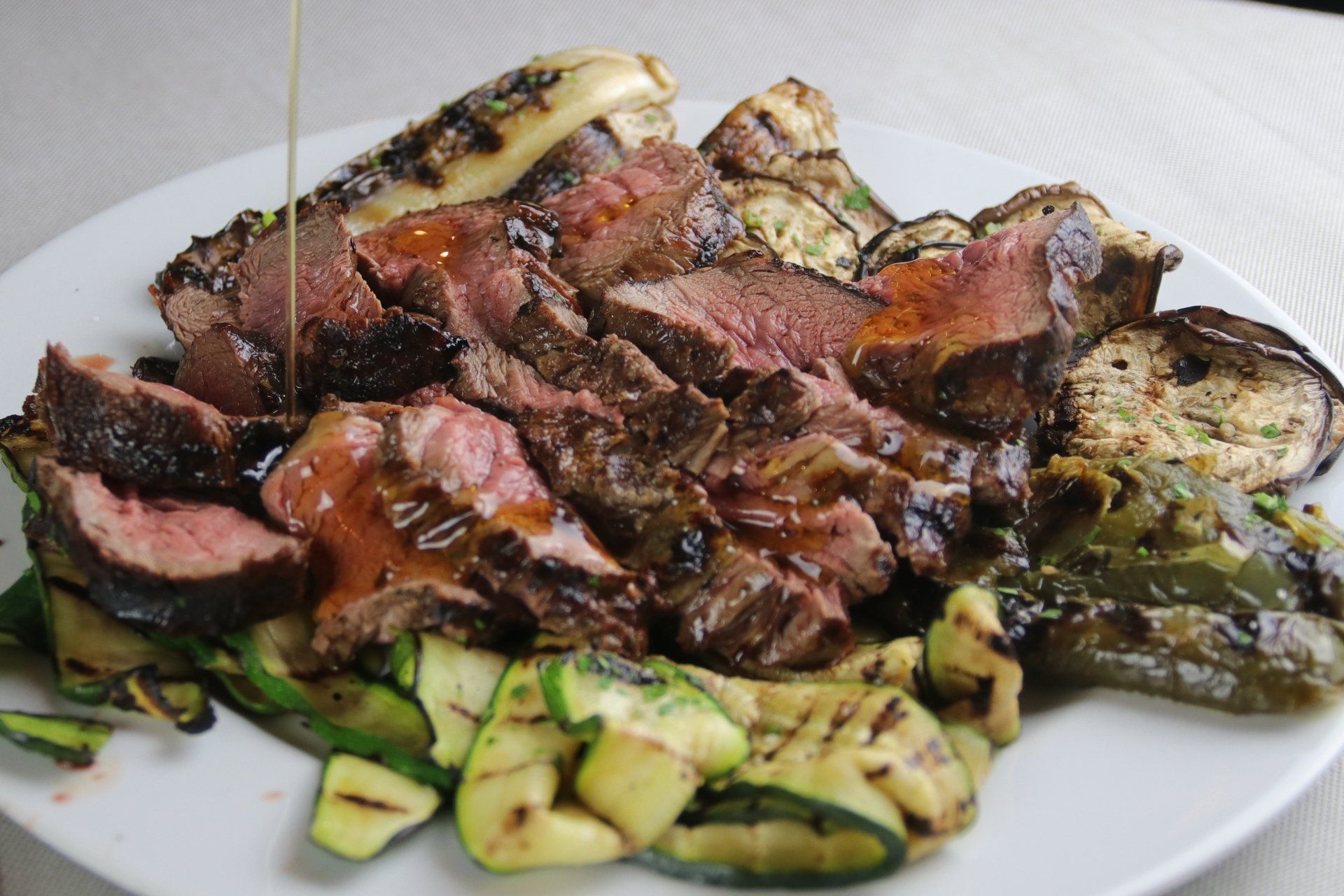 Bistecca alla griglia tagliata a fette con verdure, condita con salsa, su un piatto bianco.