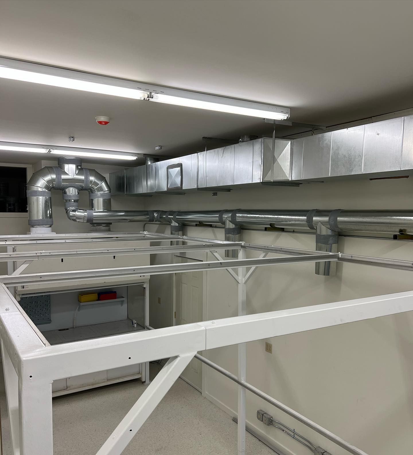 Industrial ceiling with silver ducts and fluorescent lights above a white metal structure.