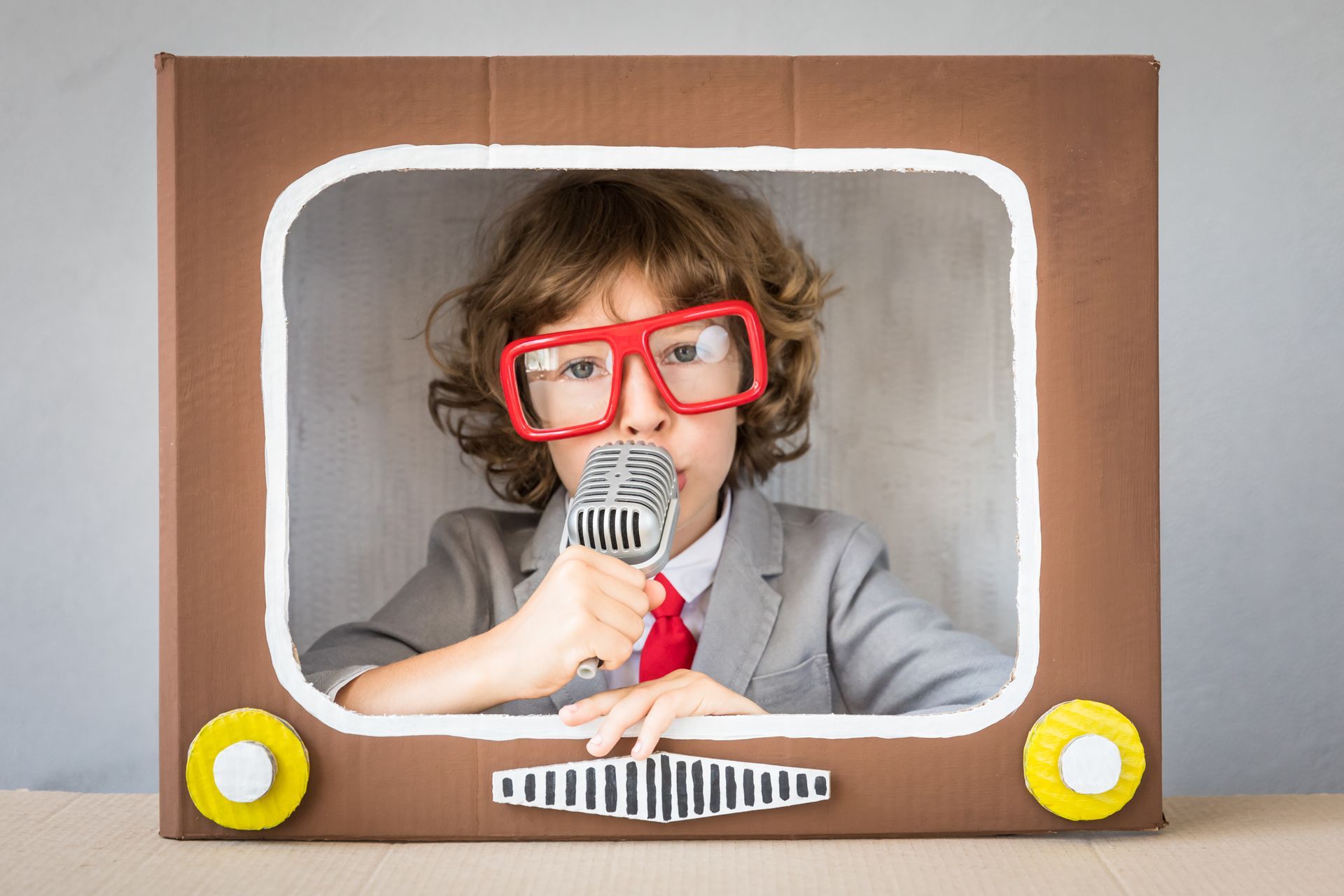 Child wearing red glasses and a gray suit sings into a toy microphone inside a cardboard TV frame.