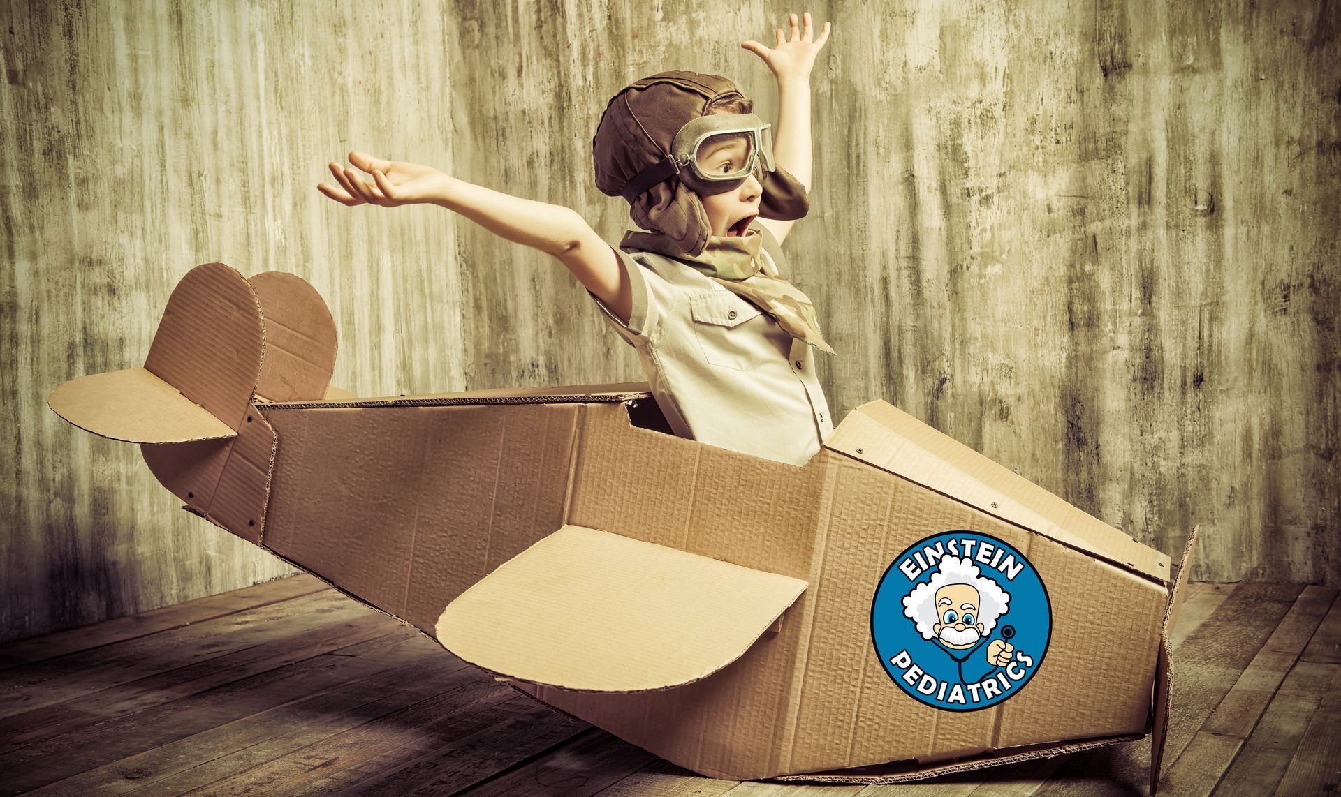 Child in cardboard airplane with arms outstretched, wearing goggles and aviator cap.