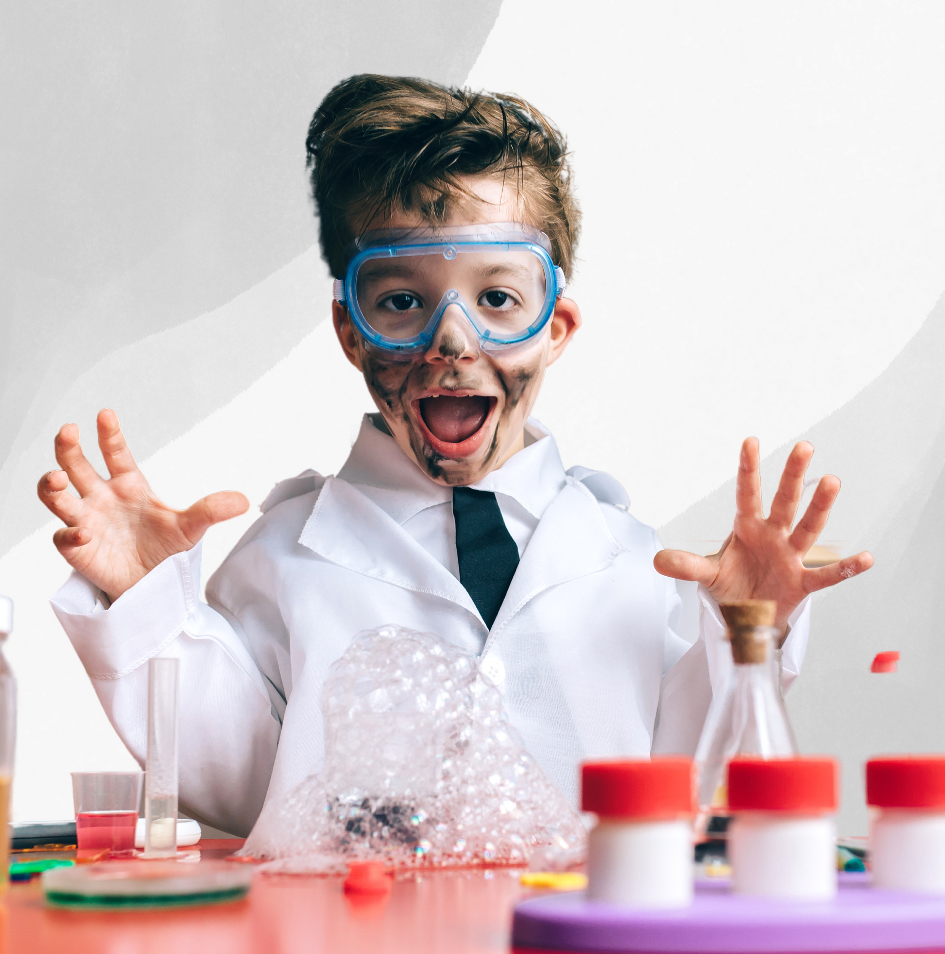 A young boy in a lab coat and goggles is sitting at a table.