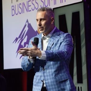 A man in a blue suit is holding a microphone in front of a business summit sign