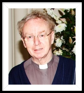 A man wearing glasses and a clergy robe is standing in front of flowers.