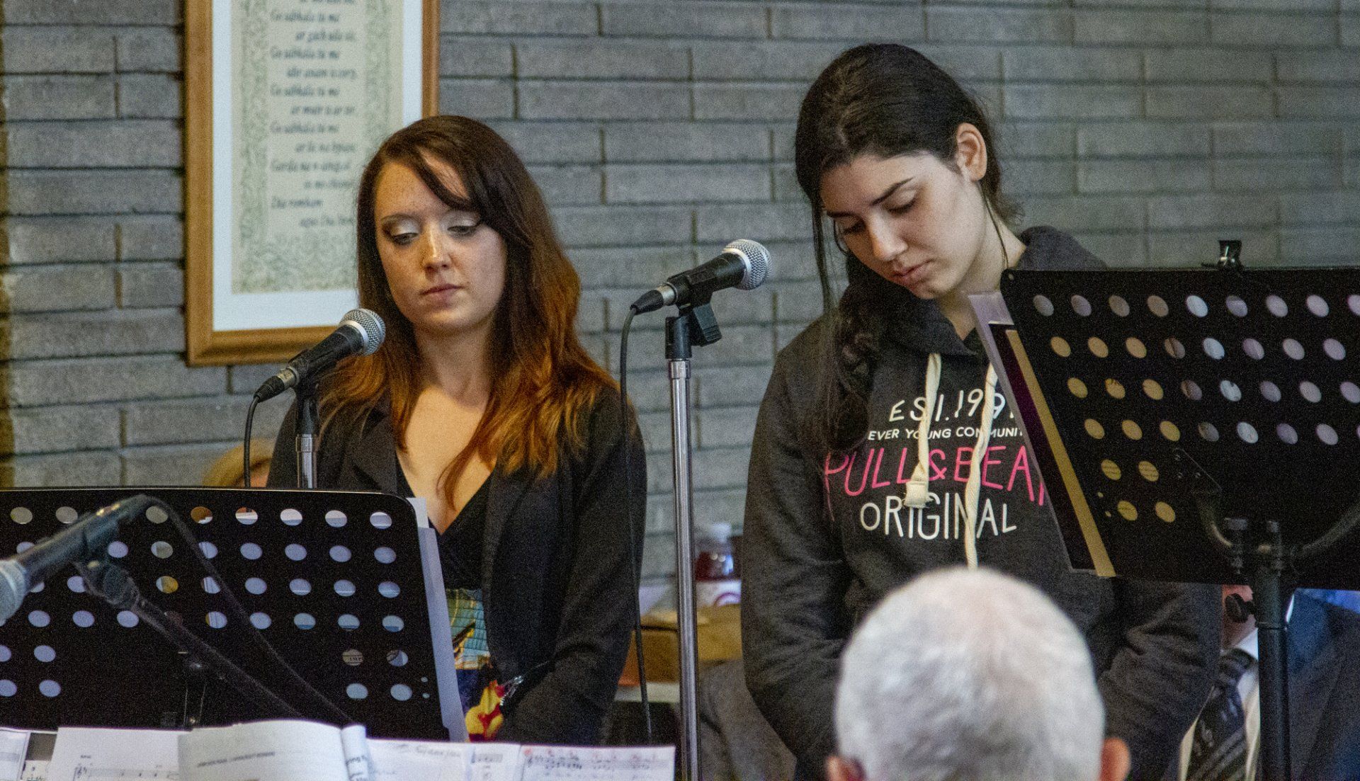 Two women are singing into microphones in front of a brick wall.
