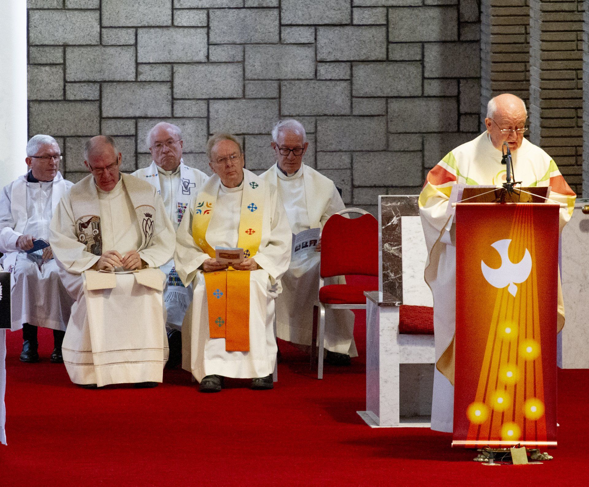 A group of priests are sitting in front of a podium with a dove on it