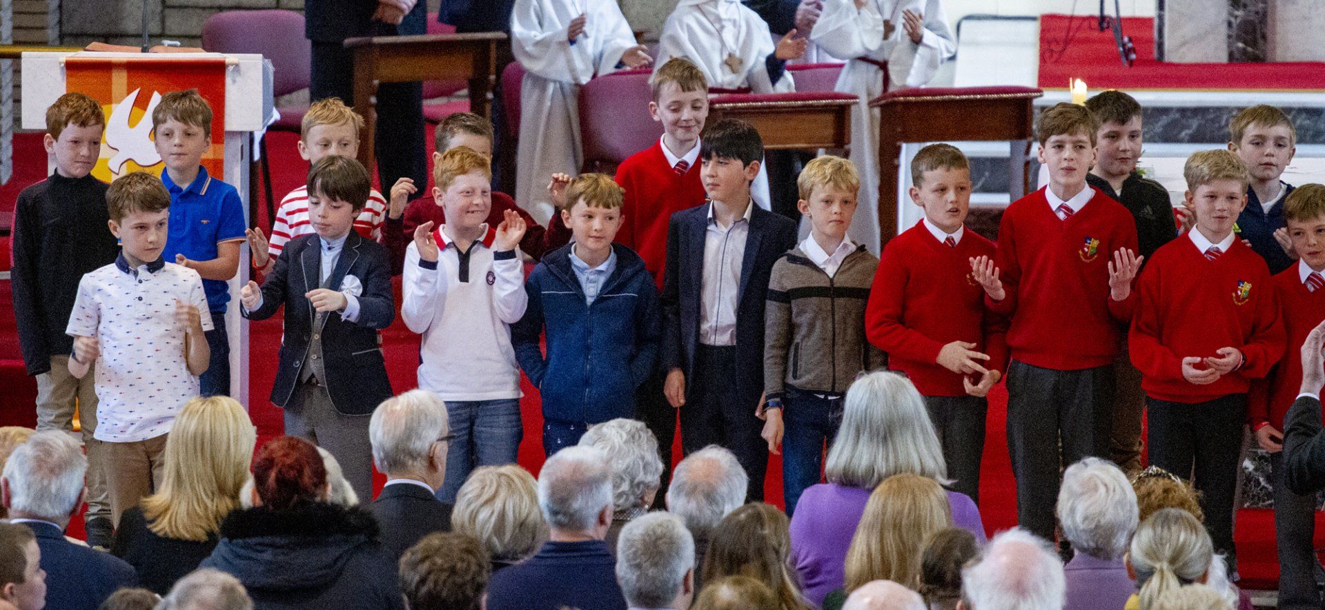 A group of children in red sweaters are standing in front of a crowd of people.