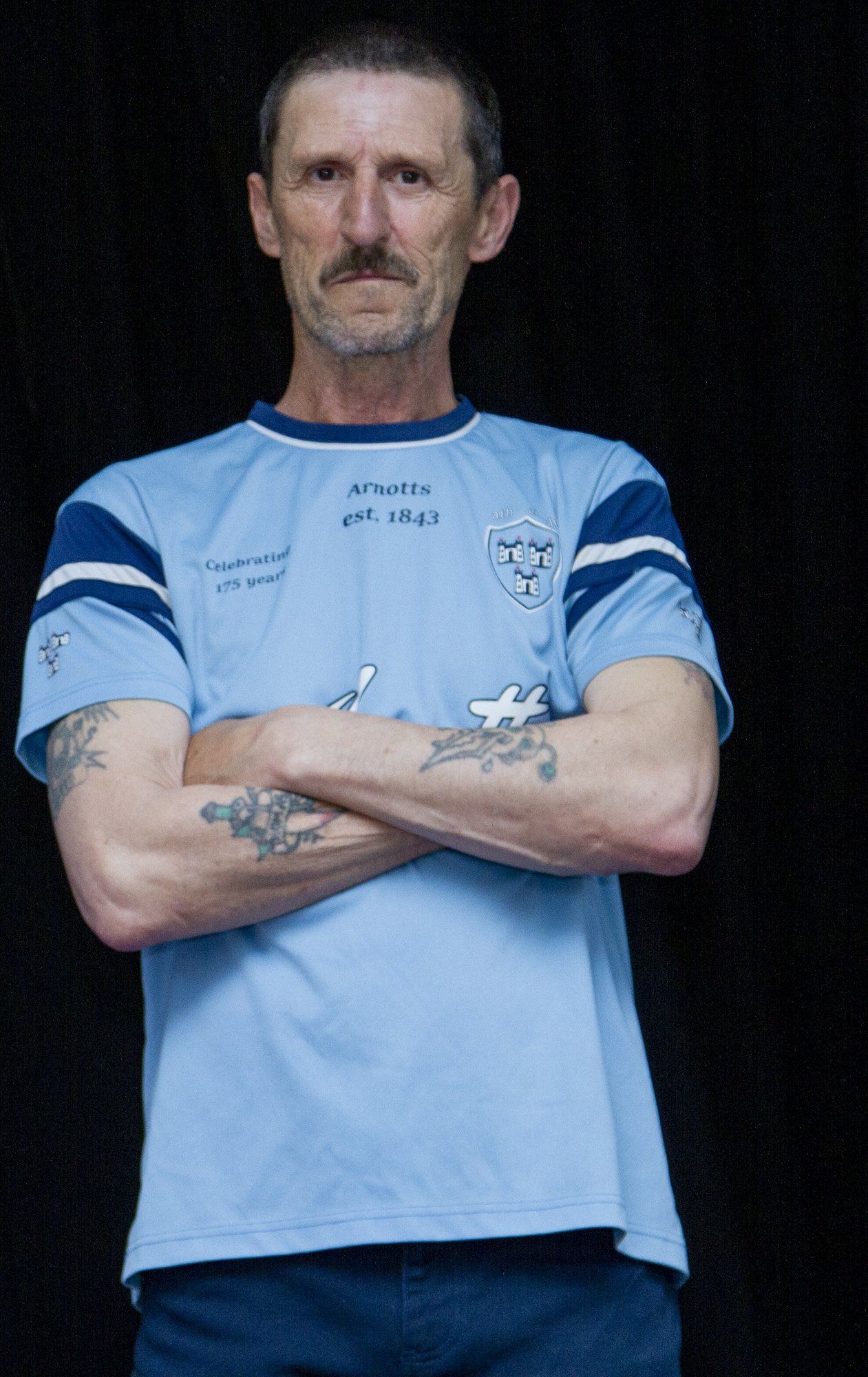 A man with a mustache is standing with his arms crossed in front of a black background.