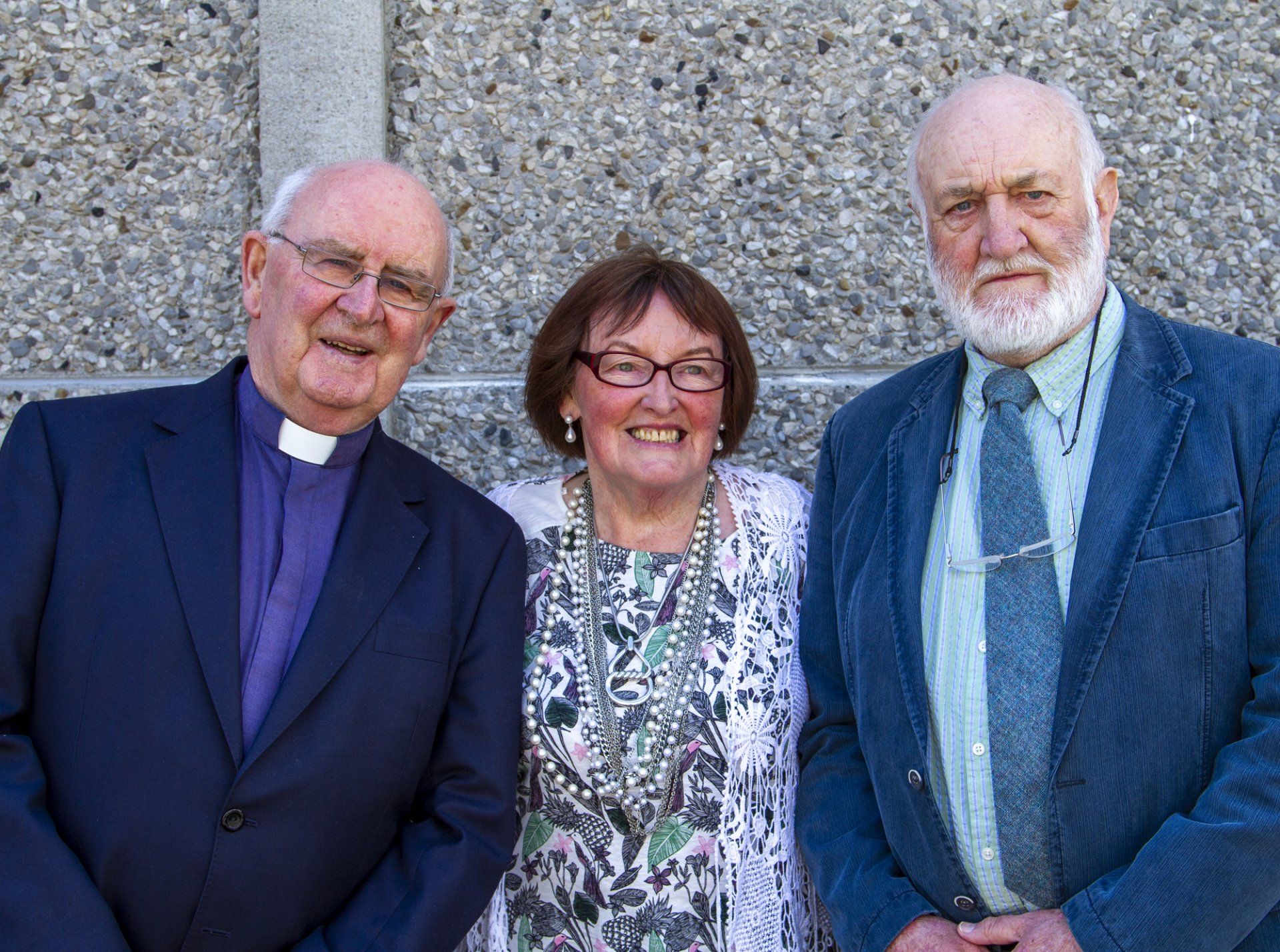 Two men and a woman are posing for a picture in front of a concrete wall.