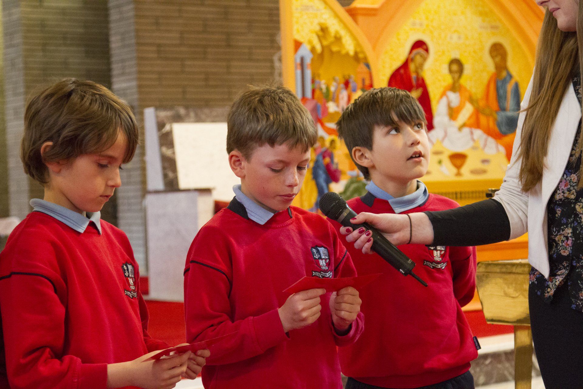 A woman is holding a microphone in front of three young boys in red sweaters.