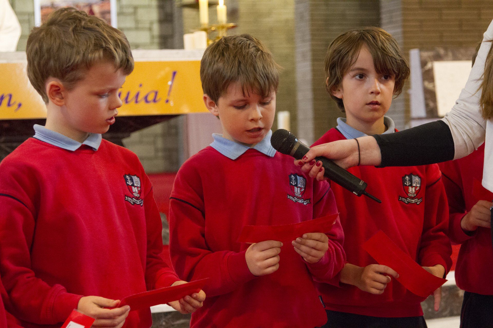 A group of young boys in red sweaters are singing into a microphone.