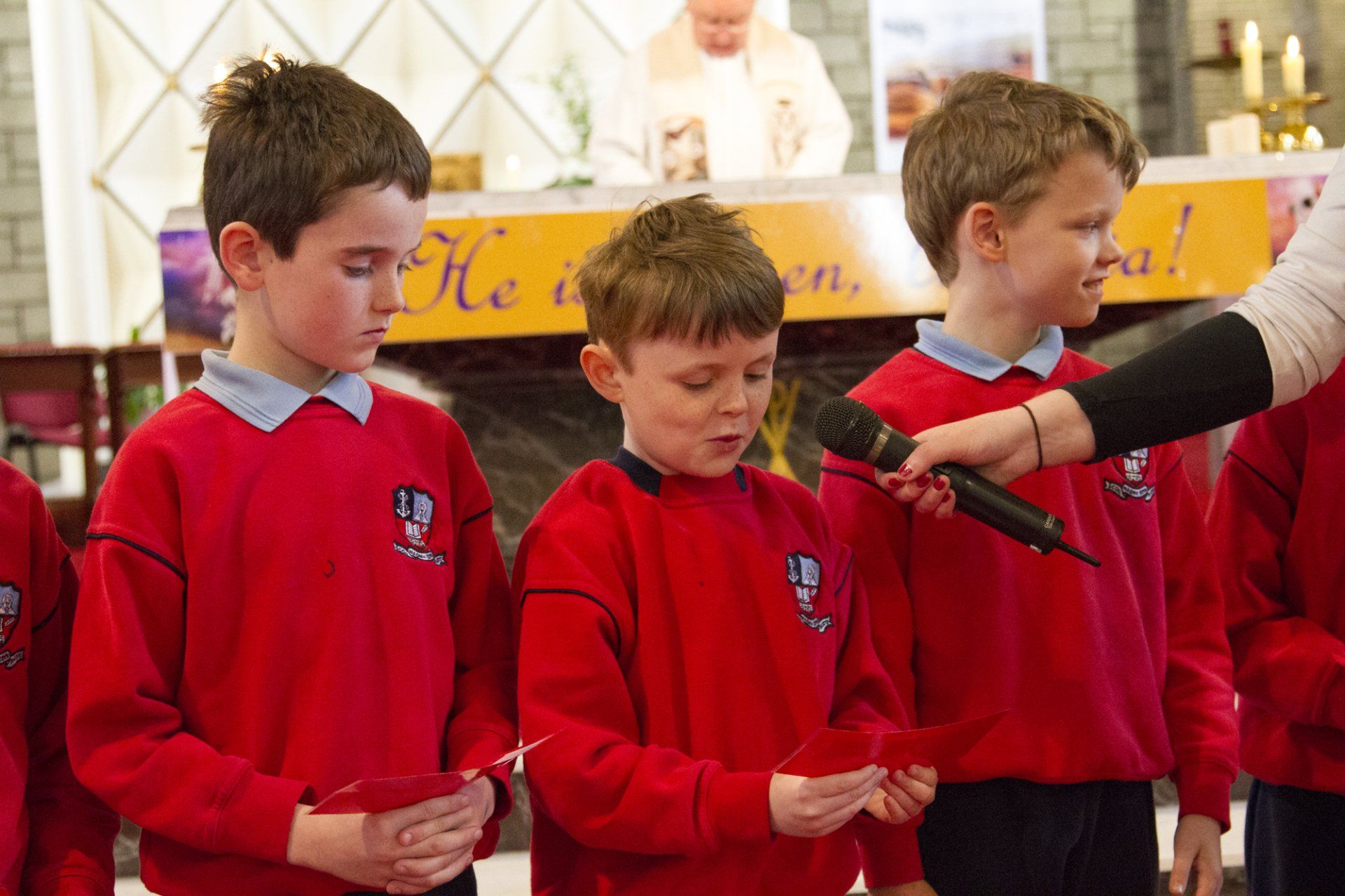 A group of young boys in red sweaters are singing into a microphone.