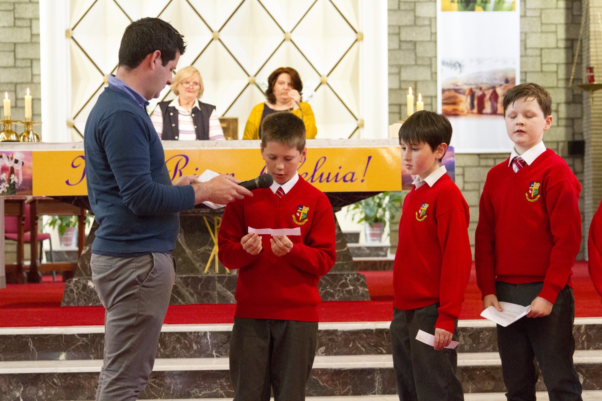 A group of young boys in red sweaters are standing in front of a man holding a piece of paper.