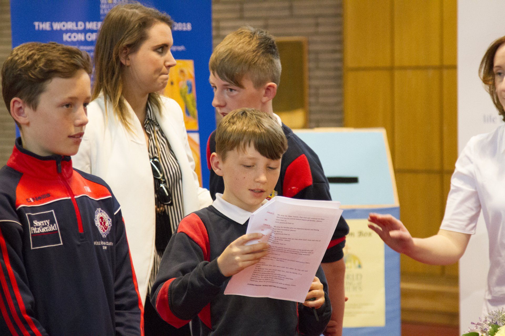 A group of children are standing around a woman holding a piece of paper.